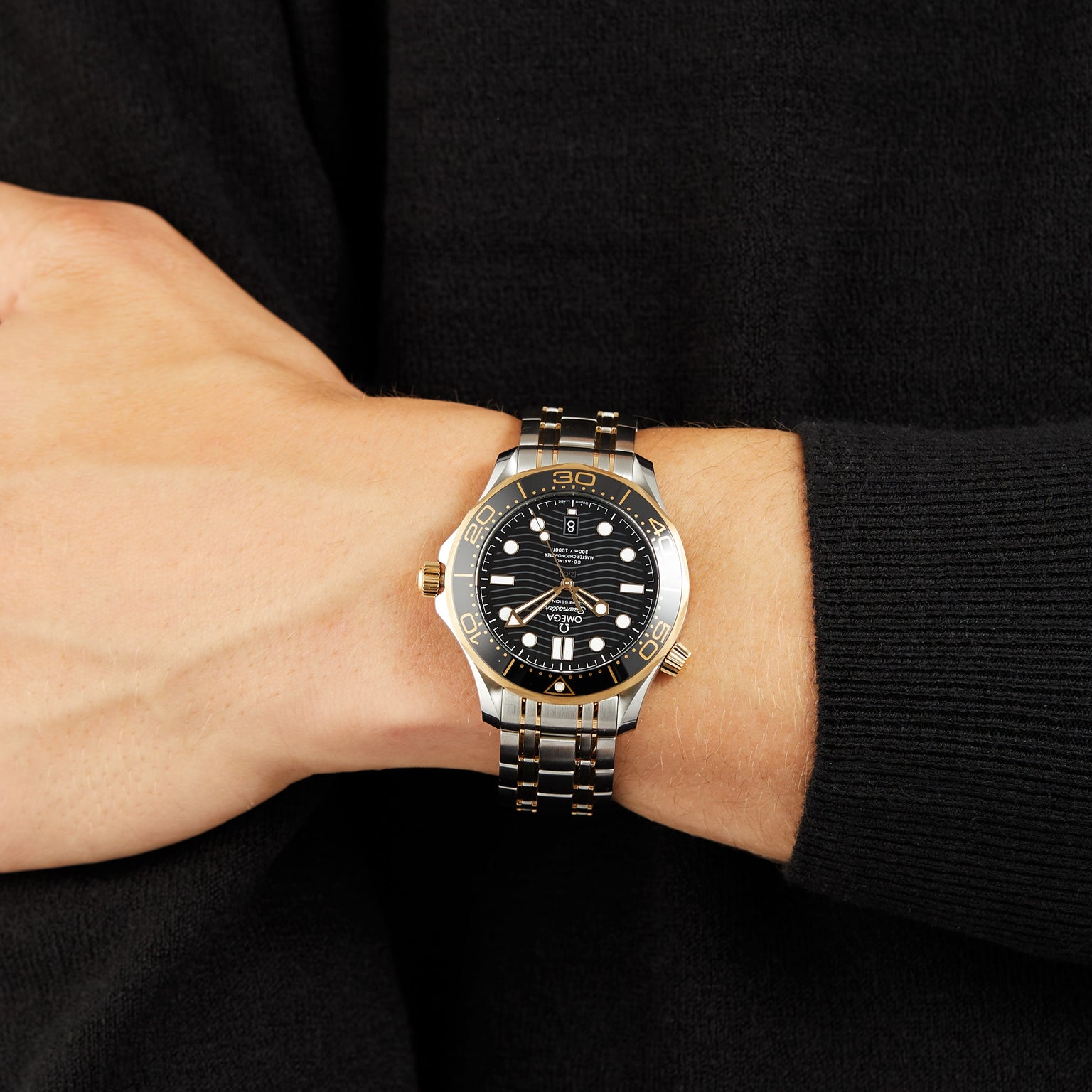 A person in a dark long-sleeve shirt wears an OMEGA Seamaster Diver 300M watch with a black dial and gold accents.