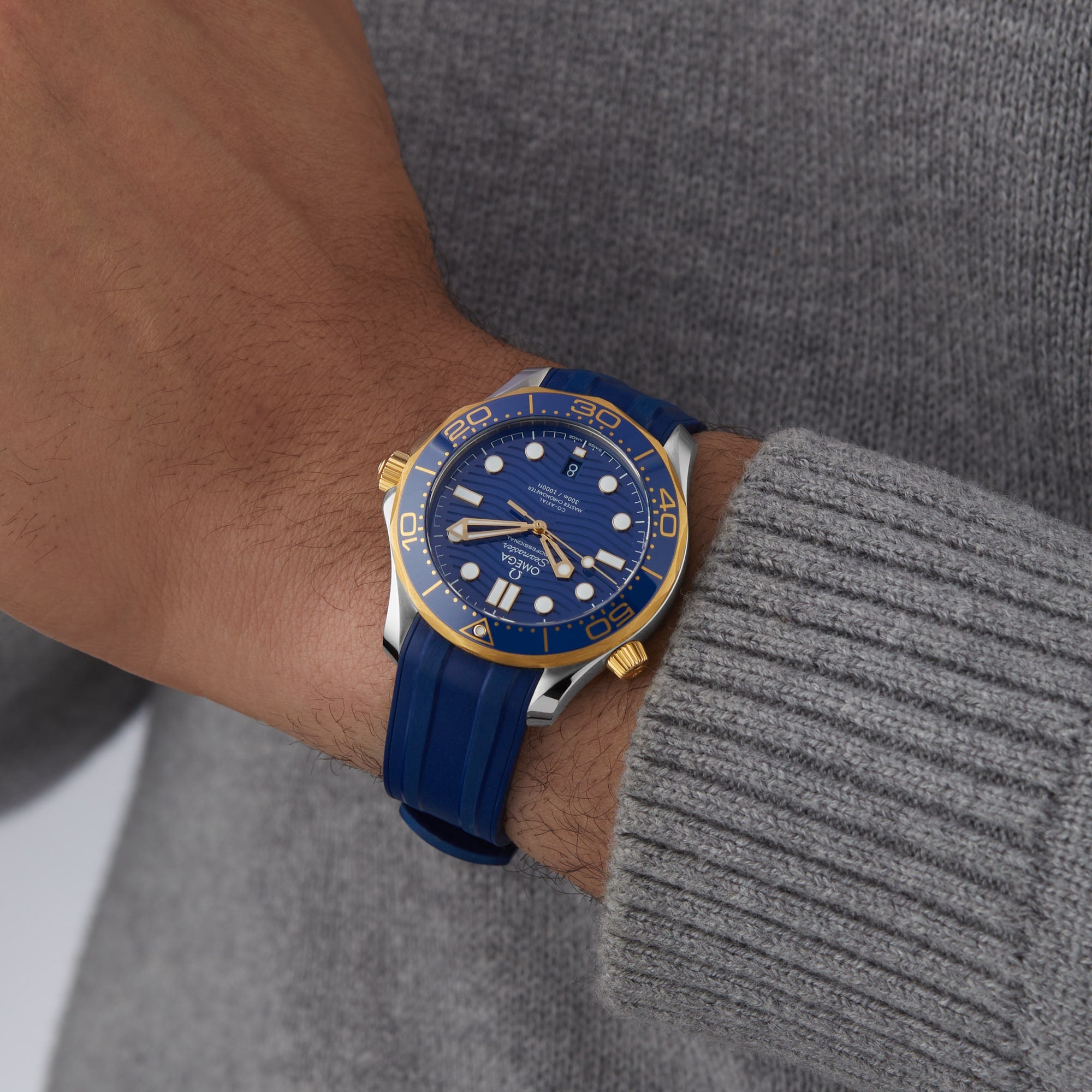 A person in a gray sweater showcases an OMEGA Seamaster Diver 300M wristwatch with a blue and gold case, blue rubber strap, blue dial, and powered by the advanced 8800 calibre movement.