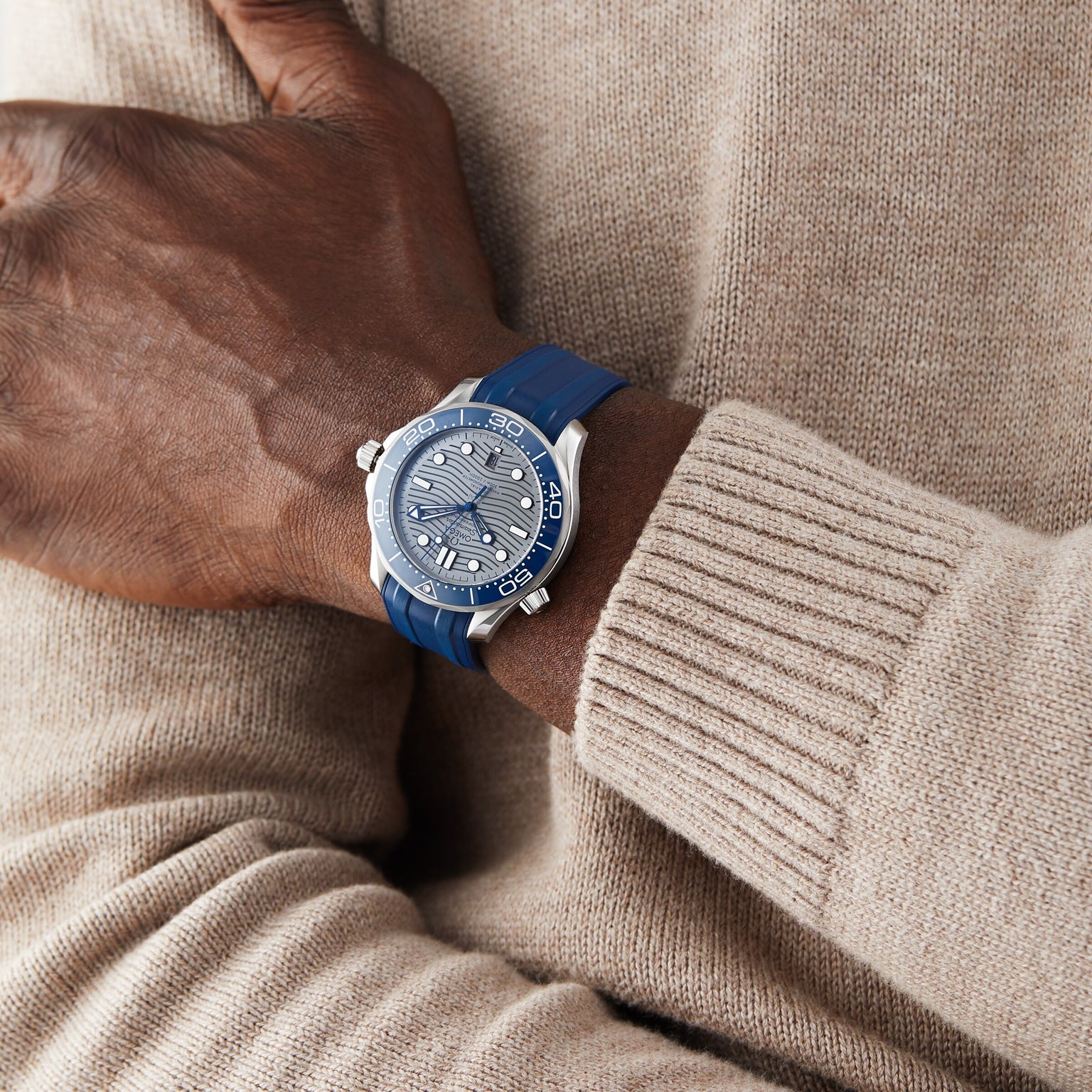 A person in a beige knit sweater showcases the OMEGA Seamaster Diver 300M, a blue and silver wristwatch with a blue strap, round detailed dial, and the precision of OMEGA Calibre 8800.