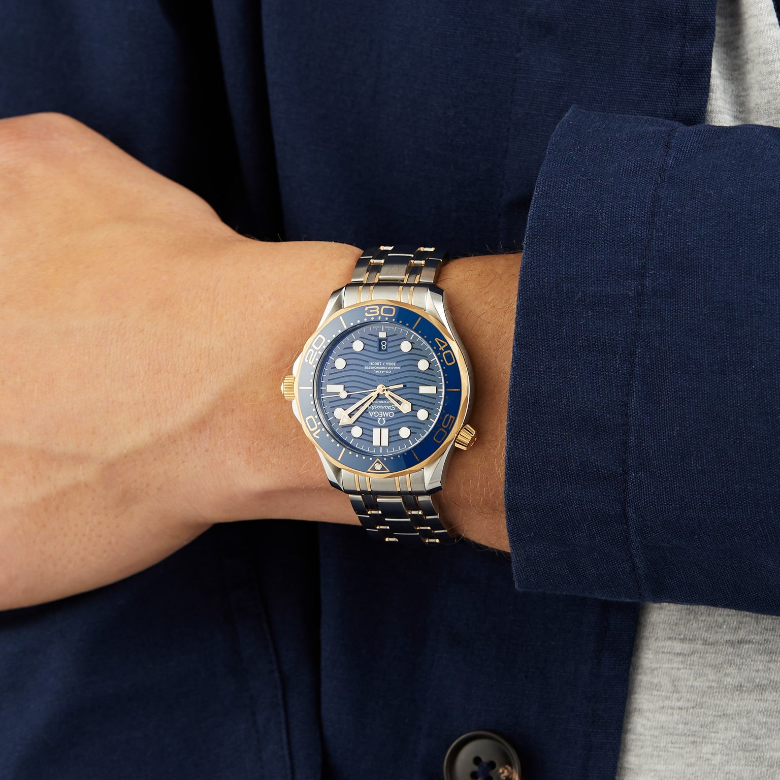 A person wearing an OMEGA Seamaster Diver 300M in blue and gold, paired with a blue jacket and gray shirt.