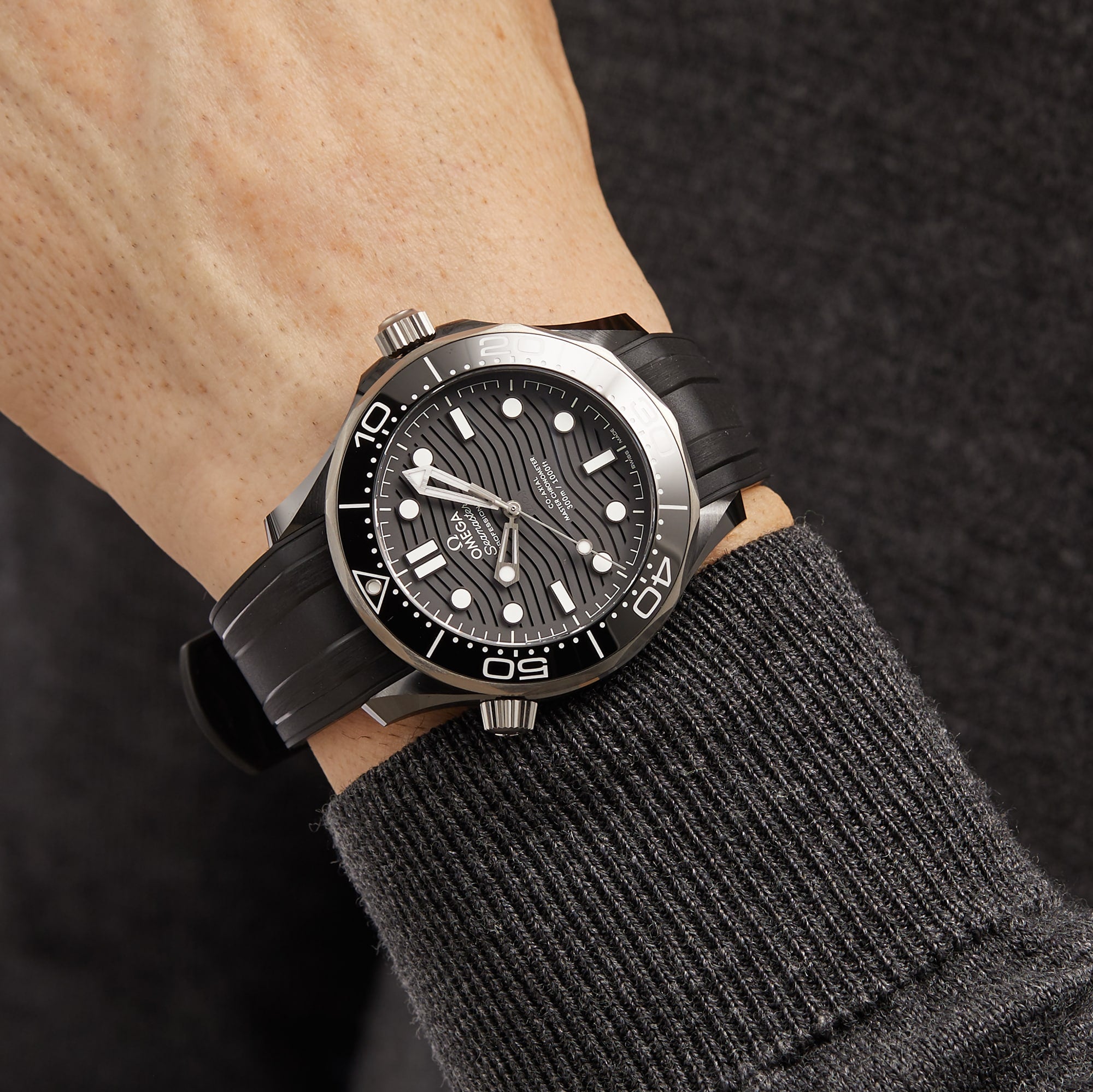 A person in a dark long-sleeve top wearing an OMEGA Seamaster Diver 300M, featuring a black and silver design, wave-patterned dial, and black rubber strap.