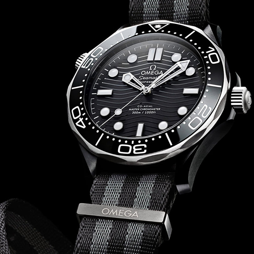 The OMEGA Seamaster Diver 300M features a black and gray NATO strap, black ceramic bezel, and wave-patterned black dial, offering Master Chronometer precision on a dark background.