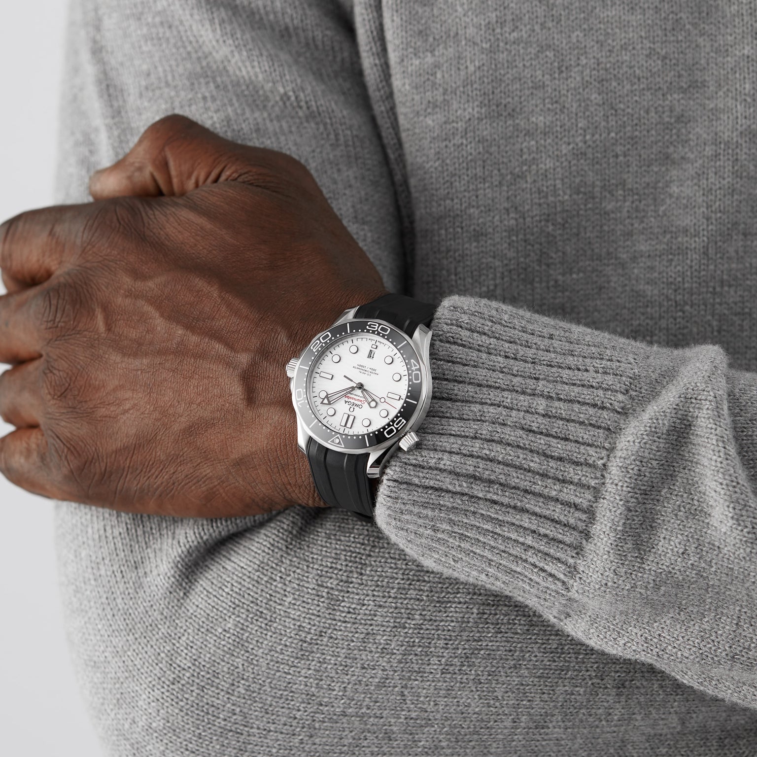 A person in a grey sweater has their arms crossed, showcasing an OMEGA Seamaster Diver 300M with a black ceramic bezel on their left wrist.