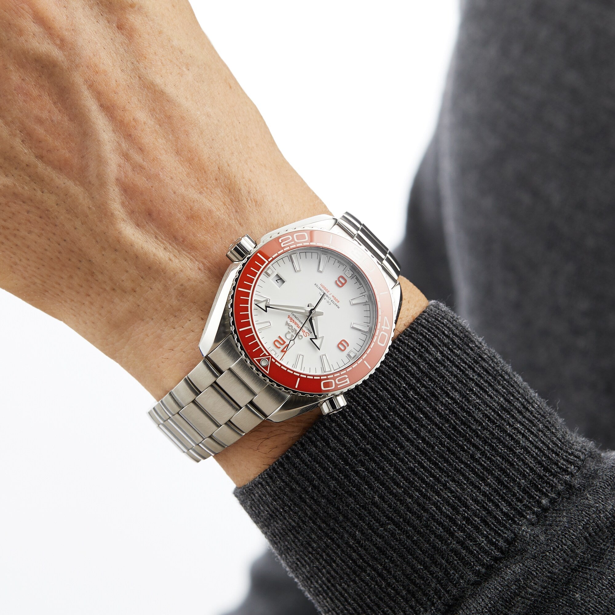 A person wearing an OMEGA Seamaster Planet Ocean 600M diving watch with a silver band, white face, and orange bezel, dressed in a dark gray long-sleeve shirt.