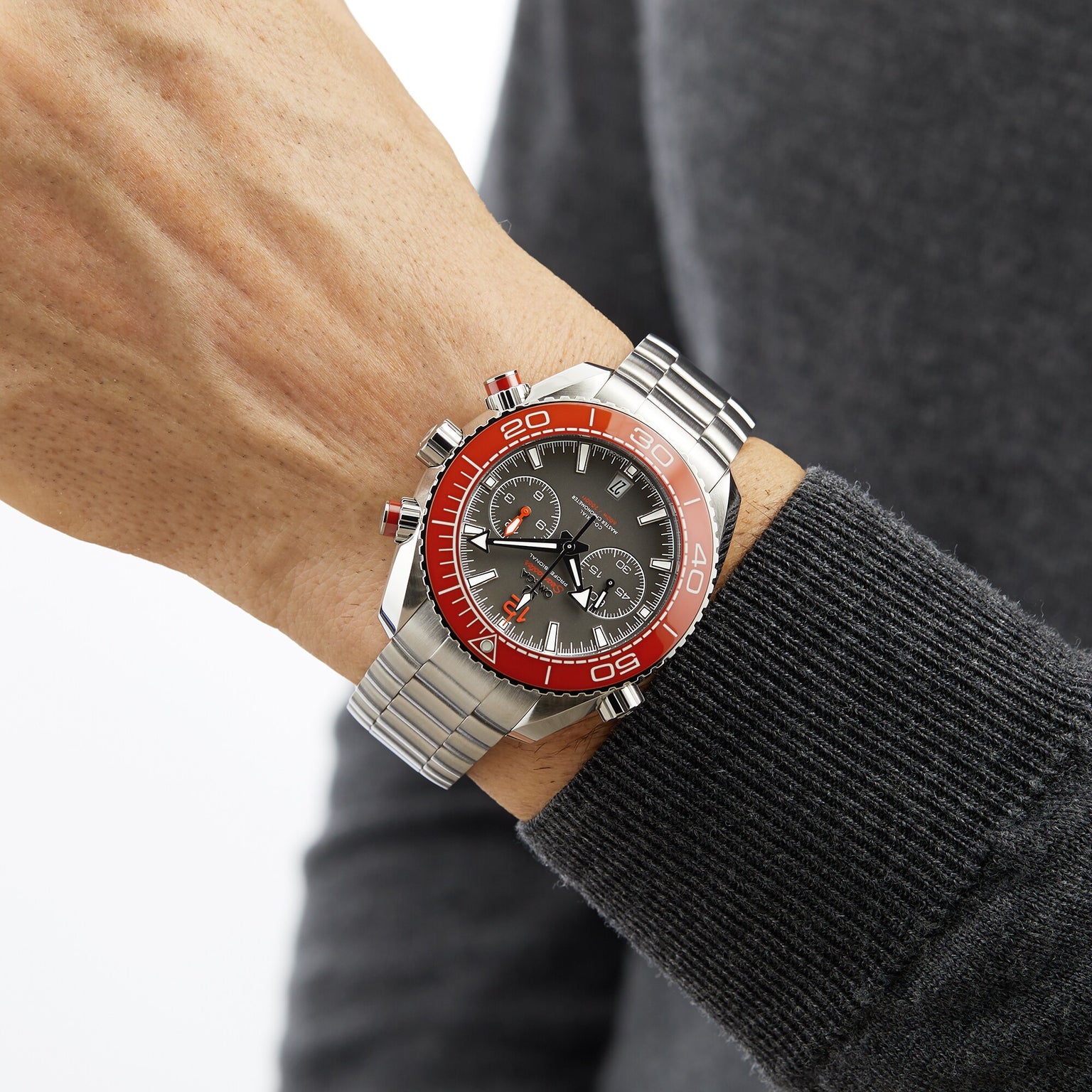 A person in a dark long-sleeve shirt wears an OMEGA Seamaster Planet Ocean 600M on their left wrist—a stainless steel watch with a red bezel, black dial, and chronograph features.