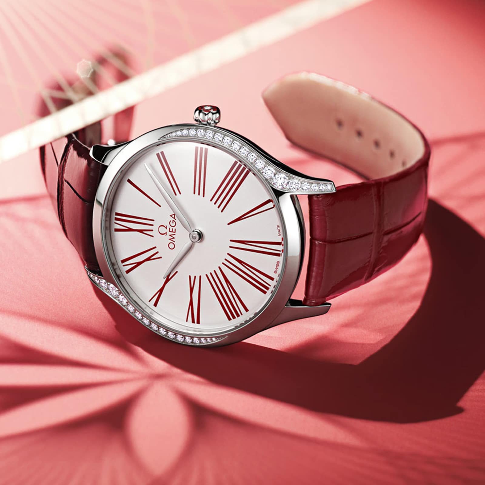 The OMEGA De Ville Tresor 36 is a diamond-accented wristwatch with a red leather strap, silver case, and white dial with bold red Roman numerals, shown on a pink surface with shadow patterns.