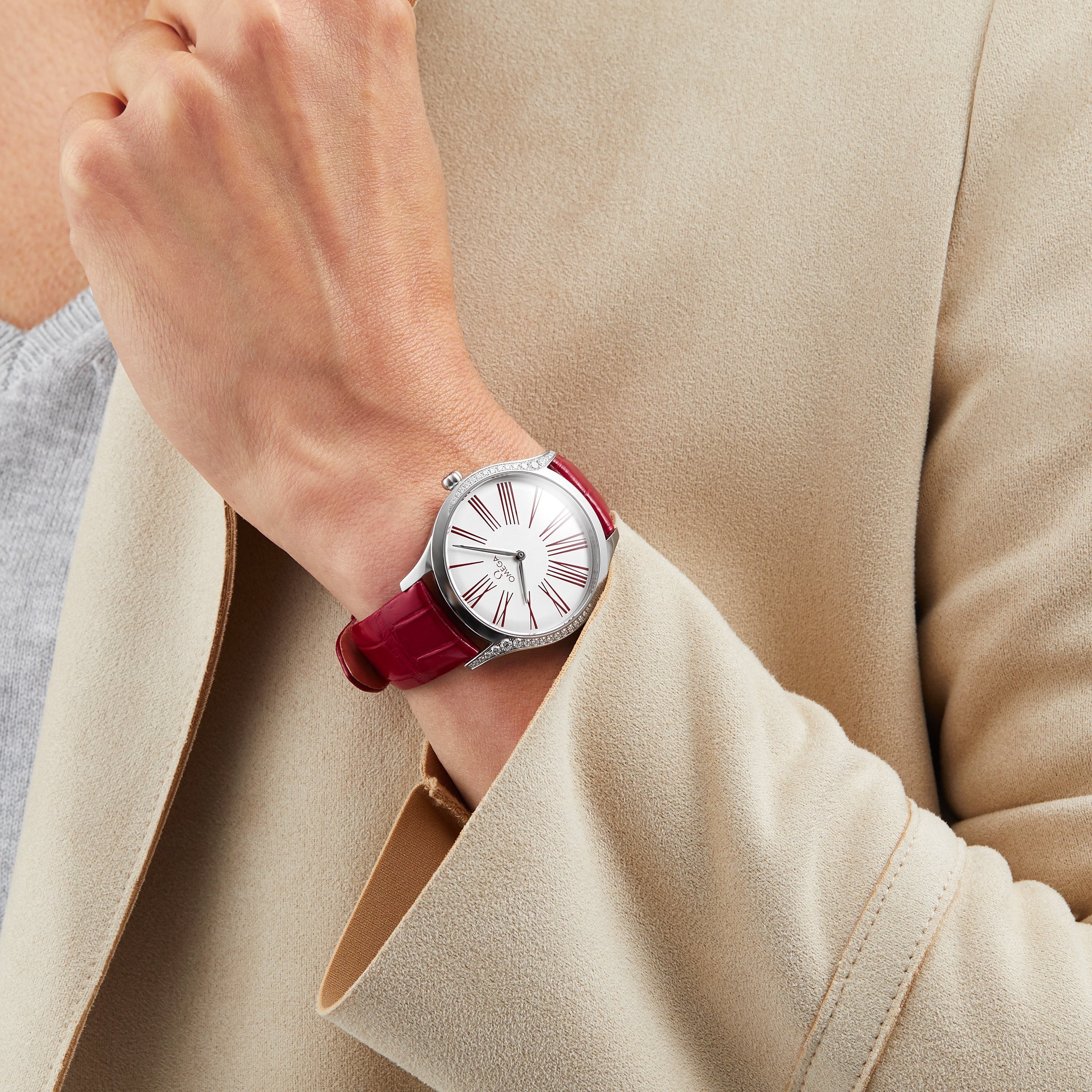 A person in a beige coat and gray top displays the OMEGA De Ville Tresor 36 wristwatch, featuring a red leather strap and white dial with bold Roman numerals.