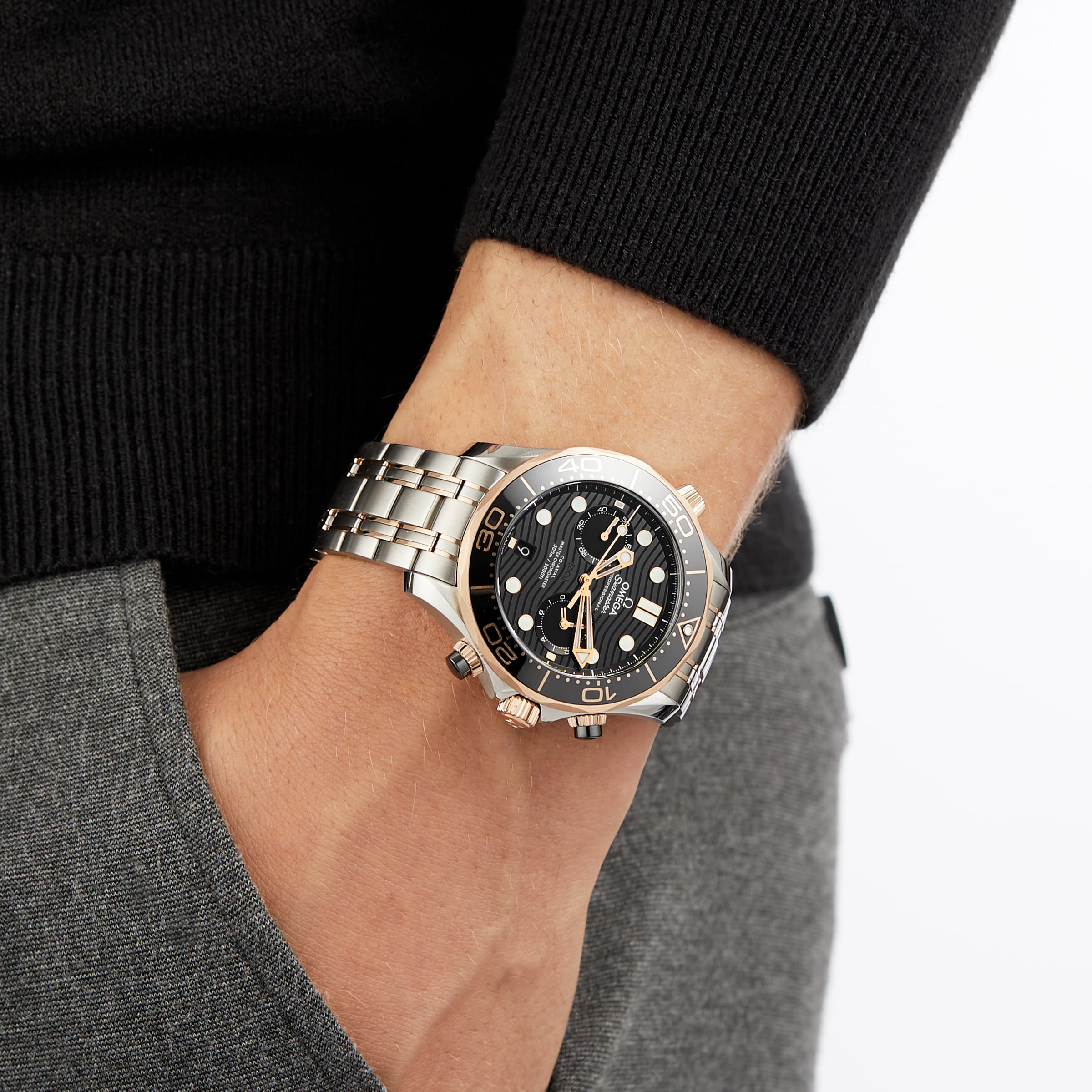 Wearing a black sweater and grey pants, a person has an OMEGA Seamaster Diver 300M with a black dial, metal band, and 18K Sedna™ gold accents on their left wrist, hand tucked in their pocket.