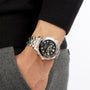 Wearing a black sweater and grey pants, a person has an OMEGA Seamaster Diver 300M with a black dial, metal band, and 18K Sedna™ gold accents on their left wrist, hand tucked in their pocket.