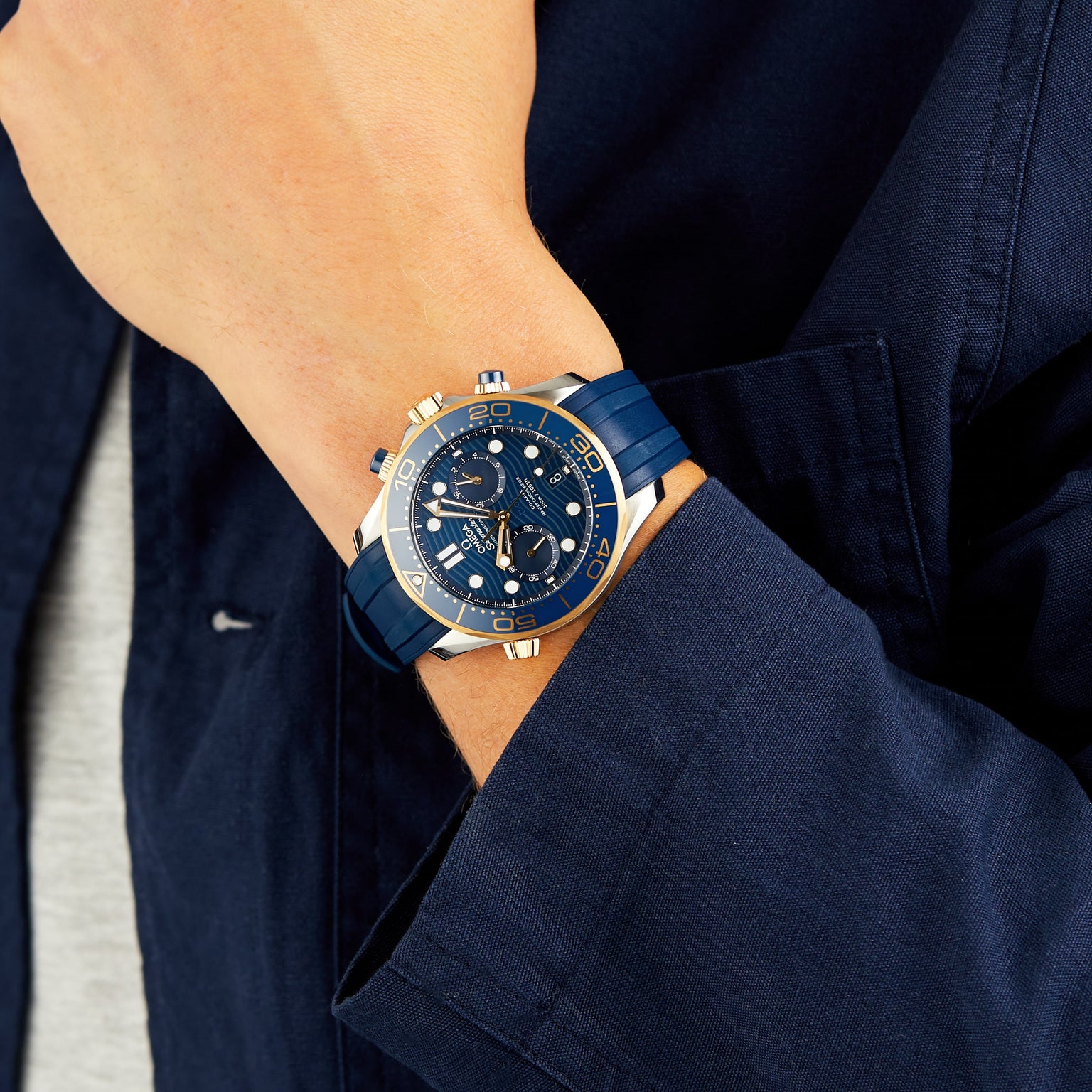 A person in a navy jacket and gray shirt wears an OMEGA Seamaster Diver 300M with a blue dial and blue strap.