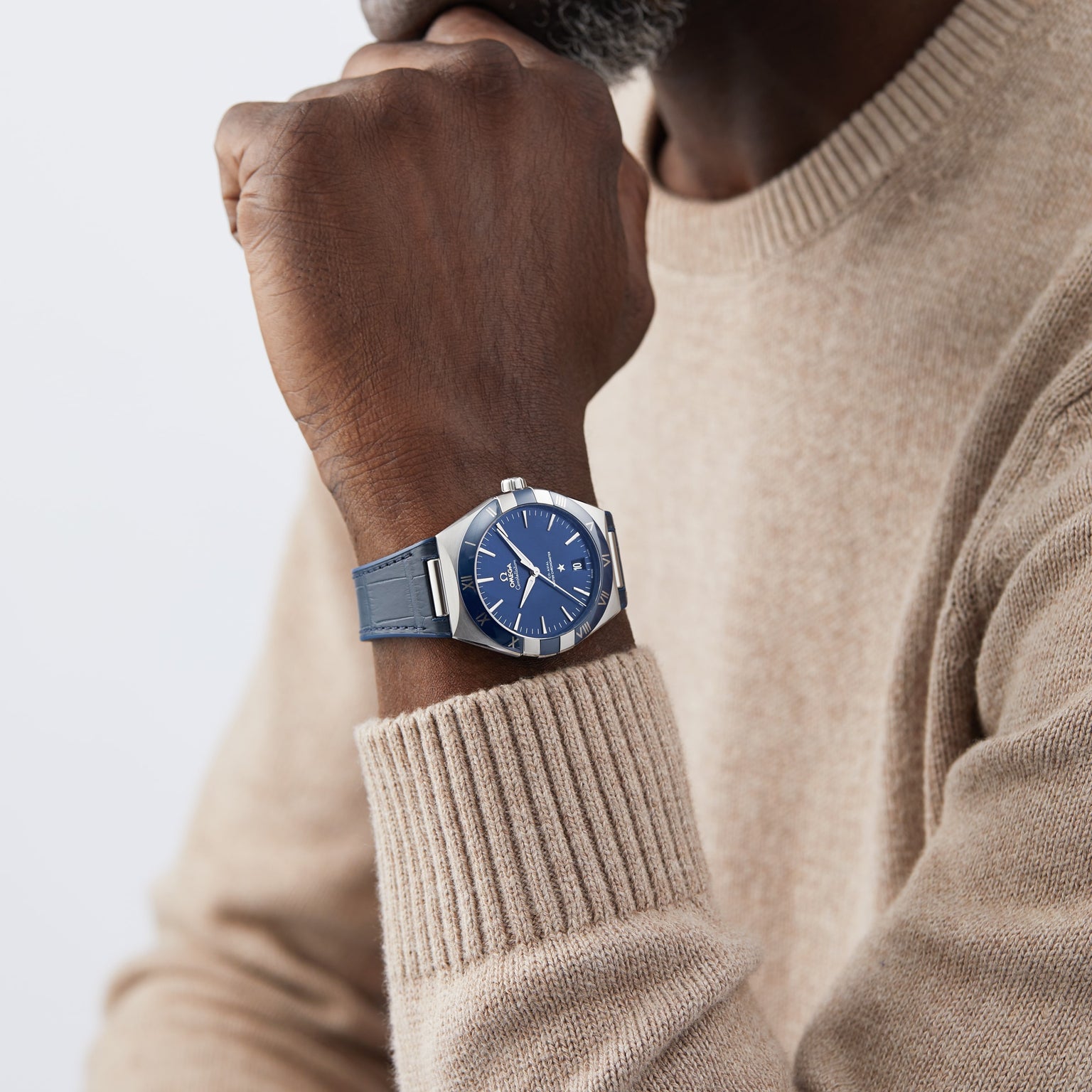 A person in a beige sweater rests their chin on their hand, showcasing the OMEGA Constellation wristwatch with a blue face and an eye-catching blue ceramic bezel.