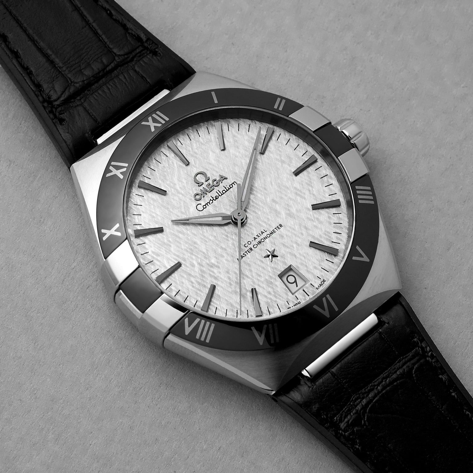A close-up of the OMEGA Constellation wristwatch shows its textured white dial, black Roman numeral bezel, date display, and black leather strap, powered by the precise 8900 Calibre movement on a light textured surface.