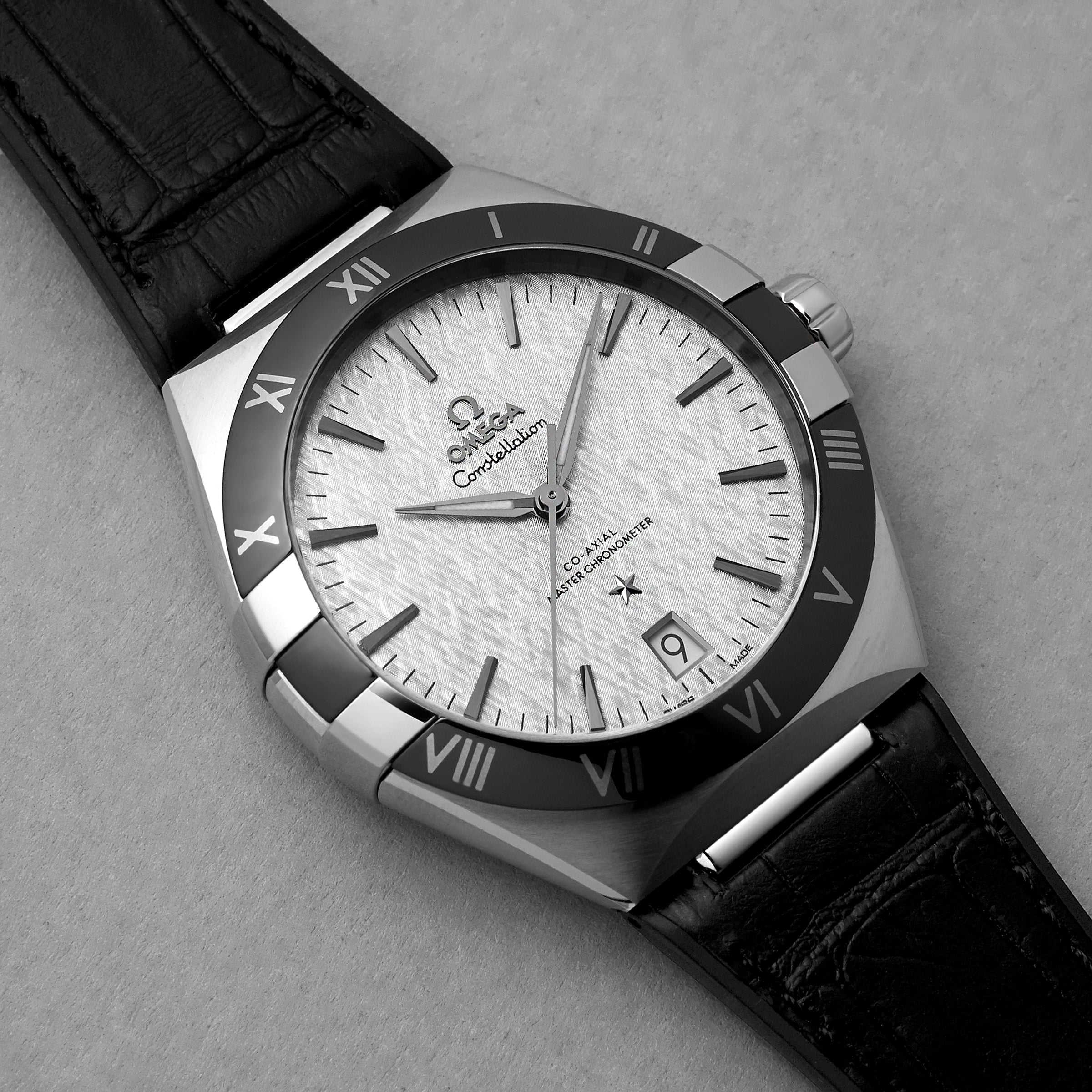 A close-up of the OMEGA Constellation wristwatch shows its textured white dial, black Roman numeral bezel, date display, and black leather strap, powered by the precise 8900 Calibre movement on a light textured surface.