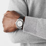 A person in a gray sweater crosses their arms, showing an OMEGA Constellation on their left wrist—a silver watch with a black band, inspired by the iconic heritage and driven by the precise 8900 calibre movement.