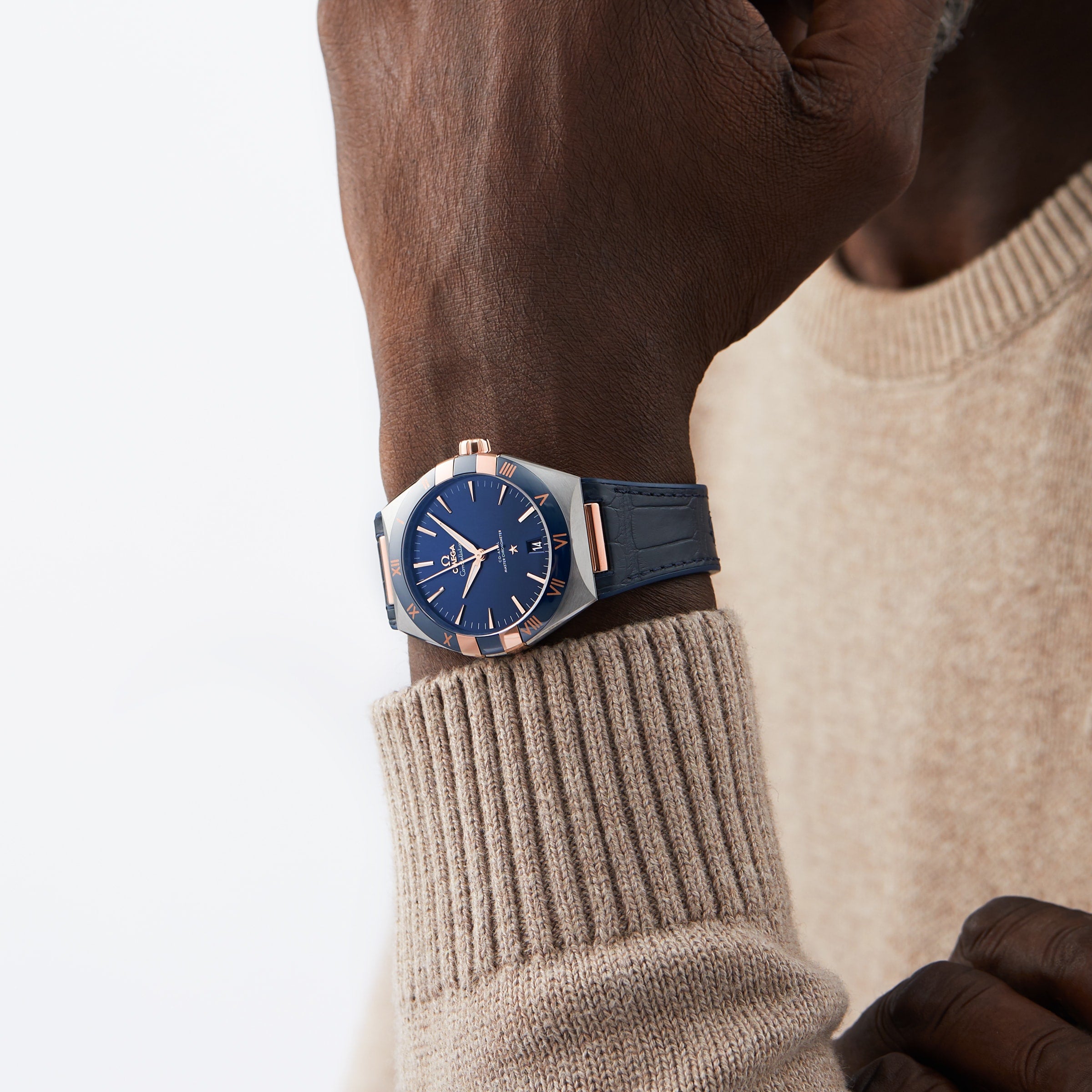 A person wearing a beige sweater showcases the OMEGA Constellation wristwatch with 8900 calibre movement and blue and rose gold design on their left wrist against a plain background.