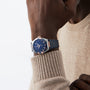 A person wearing a beige sweater showcases the OMEGA Constellation wristwatch with 8900 calibre movement and blue and rose gold design on their left wrist against a plain background.