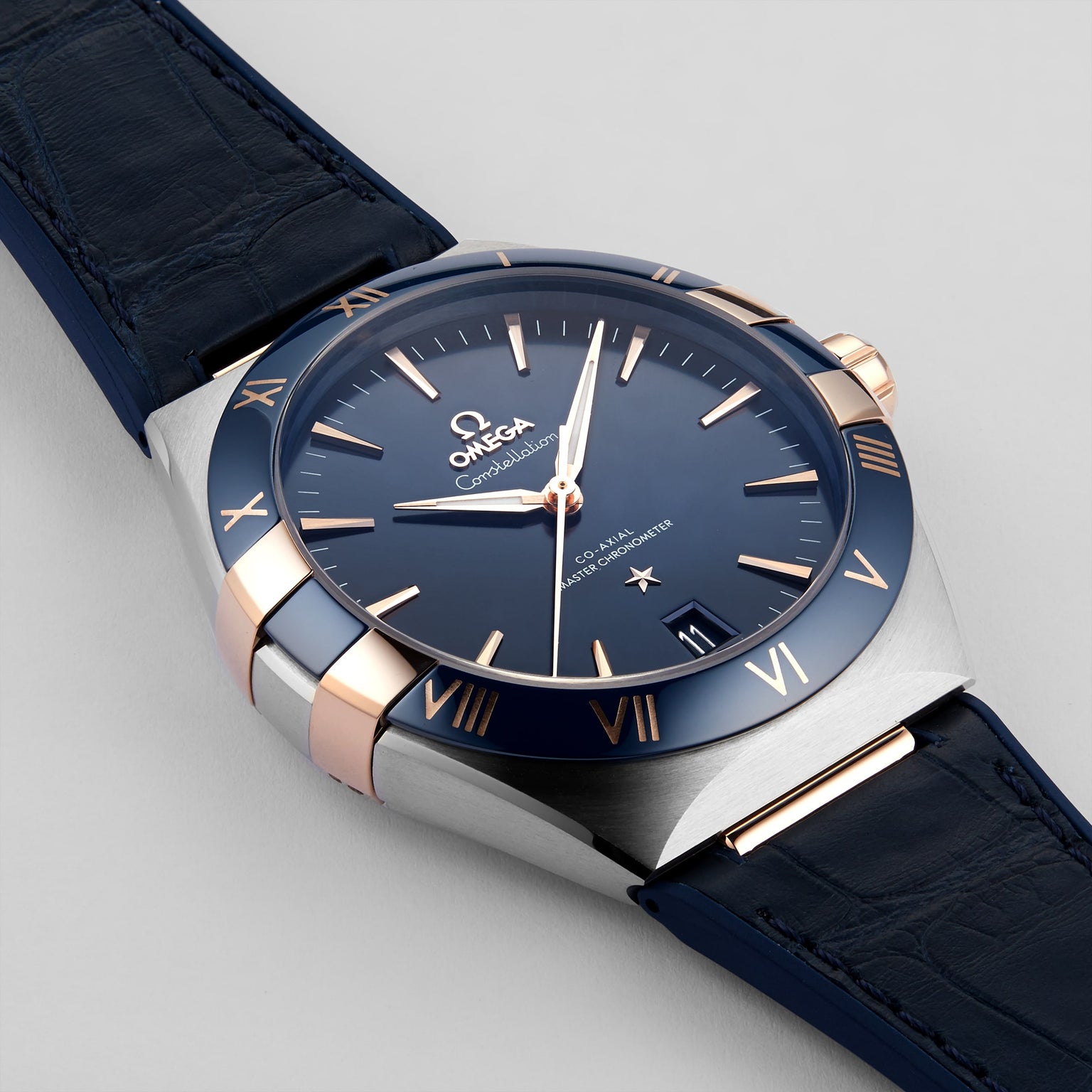 A close-up of the OMEGA Constellation wristwatch, featuring a blue dial, blue leather strap, and gold accents, showcases its Master Chronometer precision at 10:08.