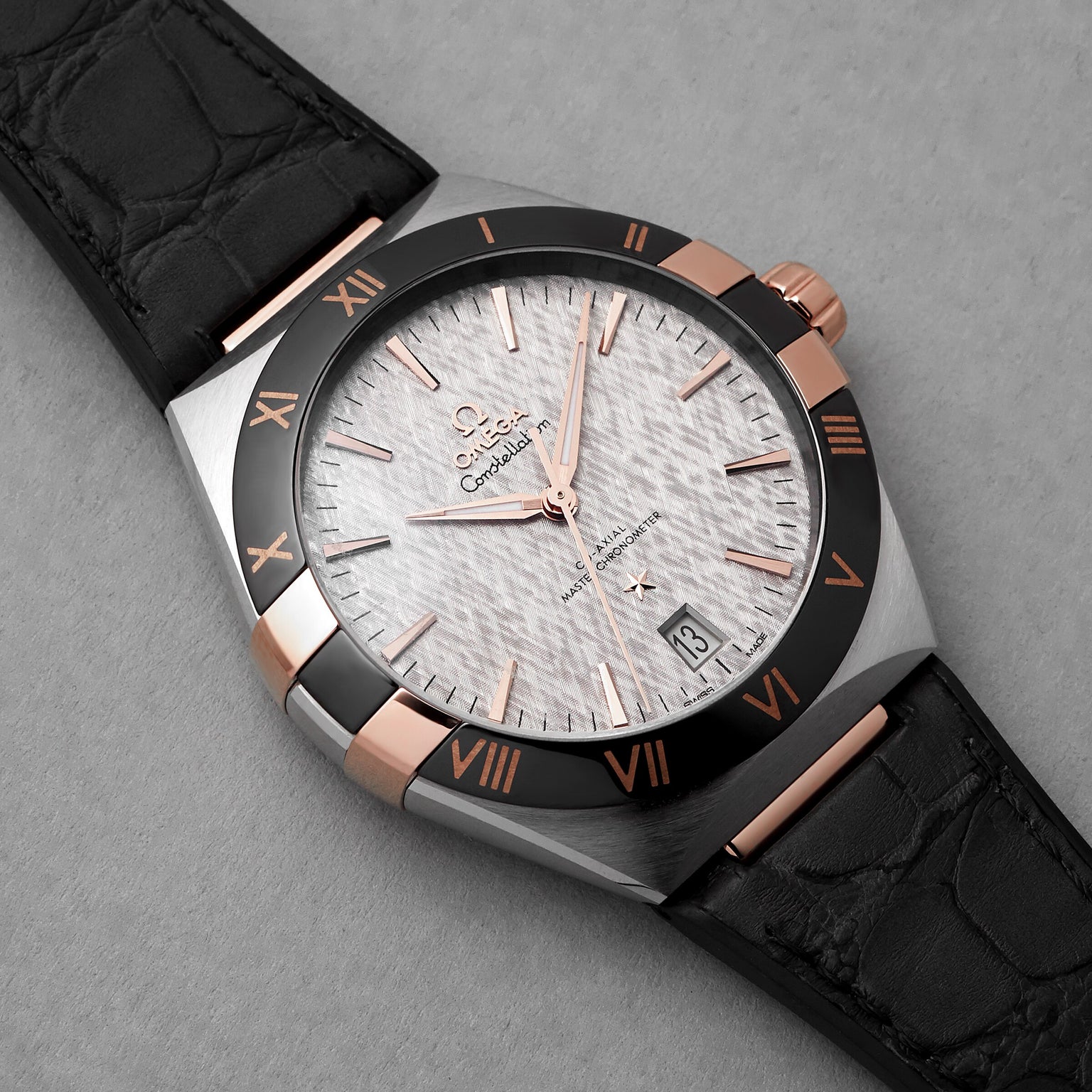 The OMEGA Constellation is a luxury wristwatch featuring a textured silver dial, black bezel with rose gold Roman numerals, black leather strap, date display at 6 o’clock, and is master chronometer certified for precision.