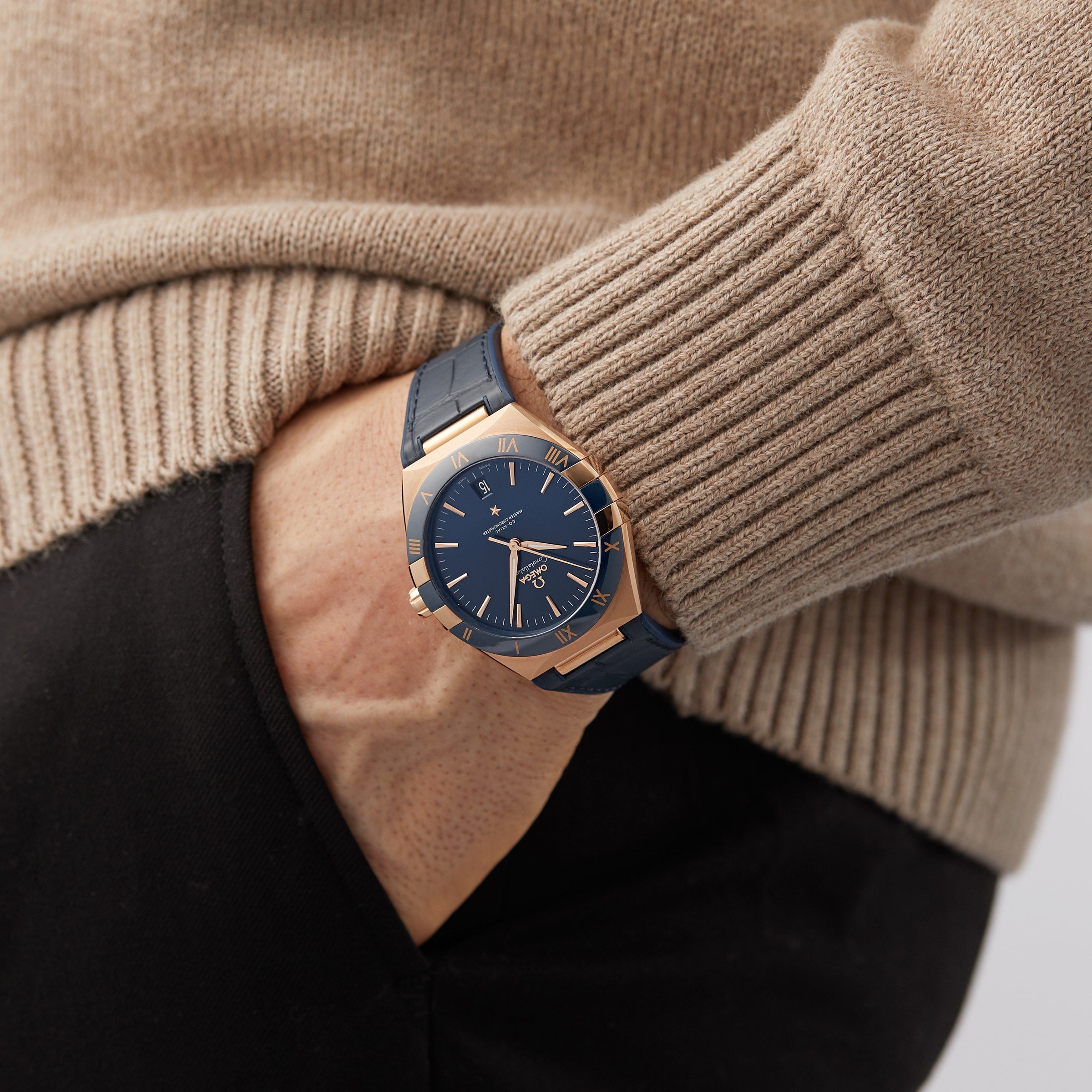 A person in a beige sweater and black pants wears an OMEGA Constellation wristwatch on their left wrist, hand partially in pocket.