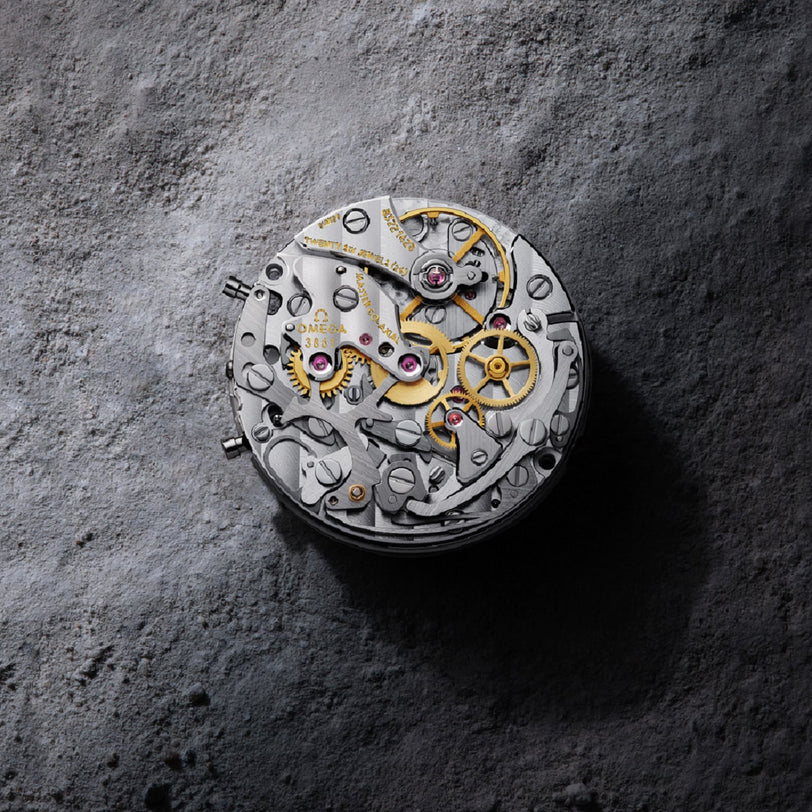 A close-up of OMEGA’s Speedmaster Moonwatch Professional reveals the intricate 3861 calibre movement with visible gears and screws, set against a textured gray surface.