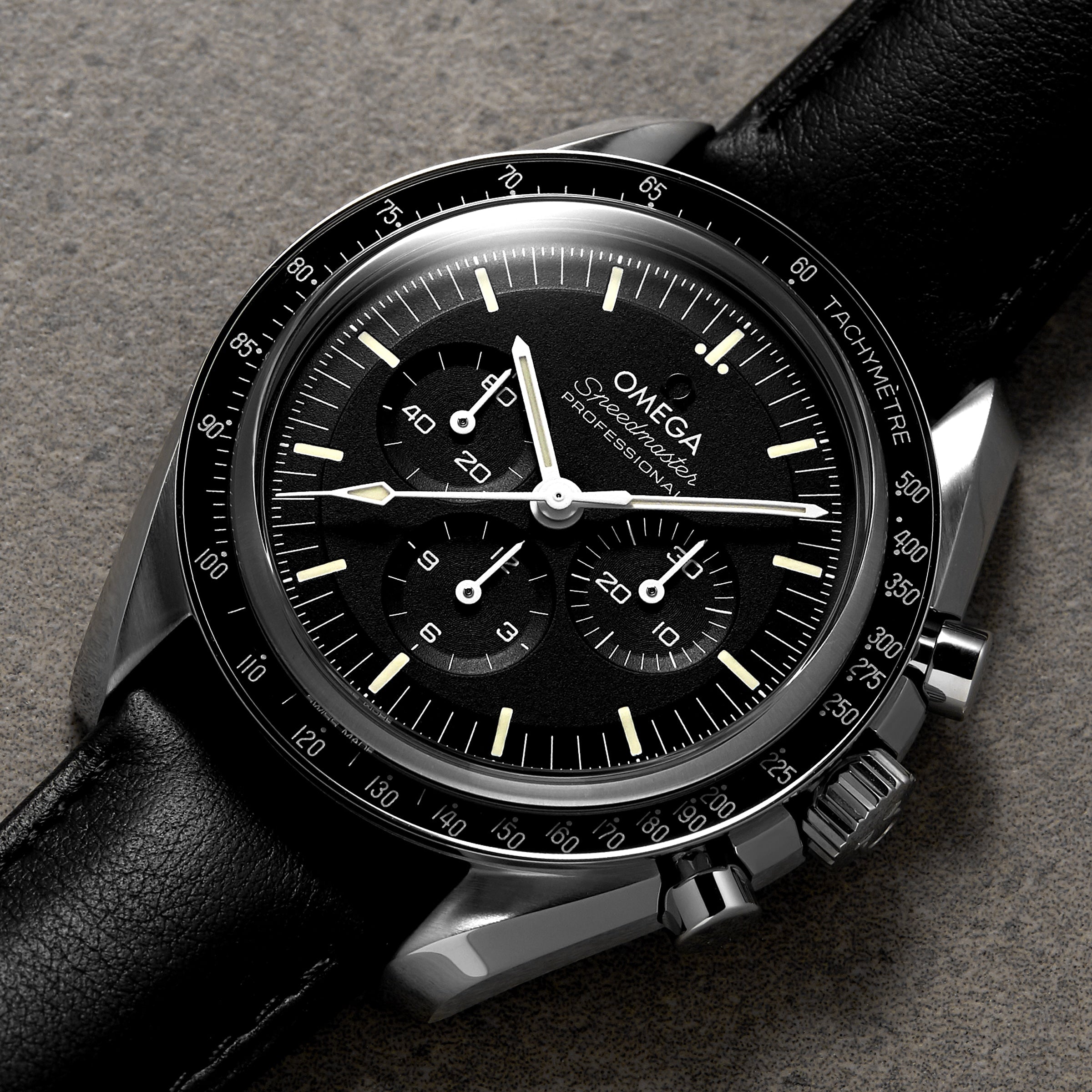 The OMEGA Speedmaster Moonwatch Professional, featuring a black dial with three sub-dials, tachymeter bezel, and black leather strap with 3861 calibre movement, is photographed on a textured gray surface.