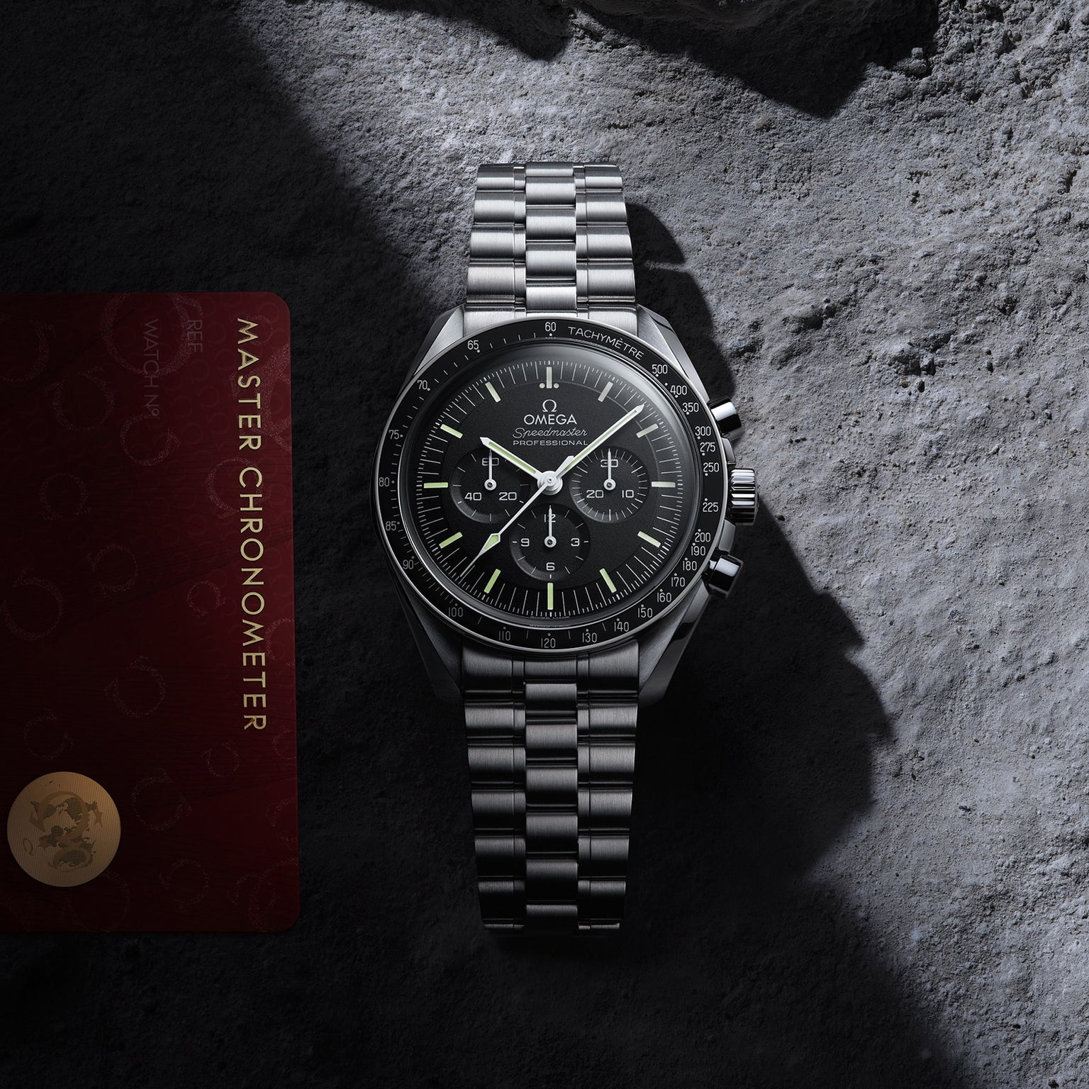 An OMEGA Speedmaster Moonwatch Professional with a black dial sits on a textured gray surface next to its red Master Chronometer certification card.