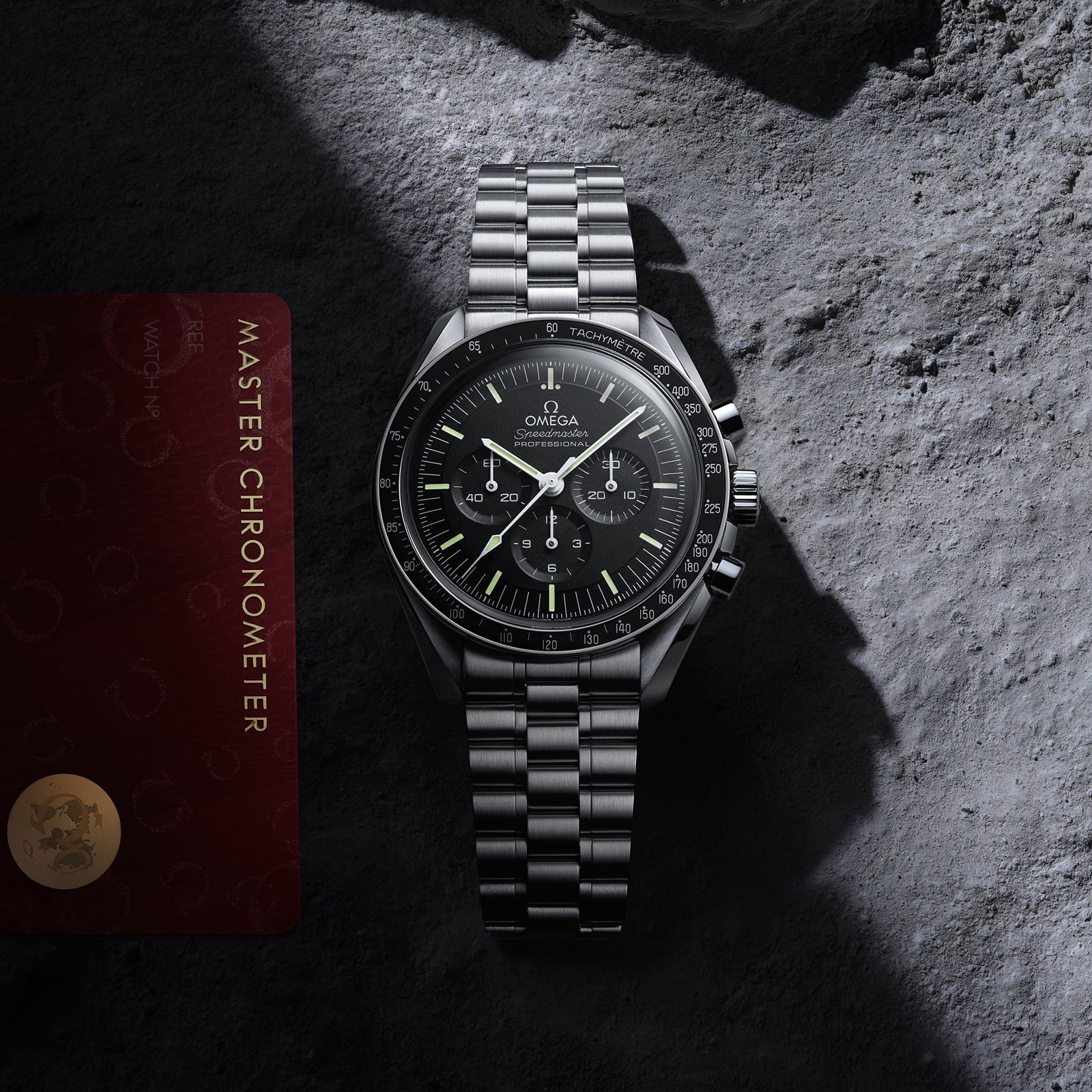 An OMEGA Speedmaster Moonwatch Professional with a black dial sits on a textured gray surface next to its red Master Chronometer certification card.