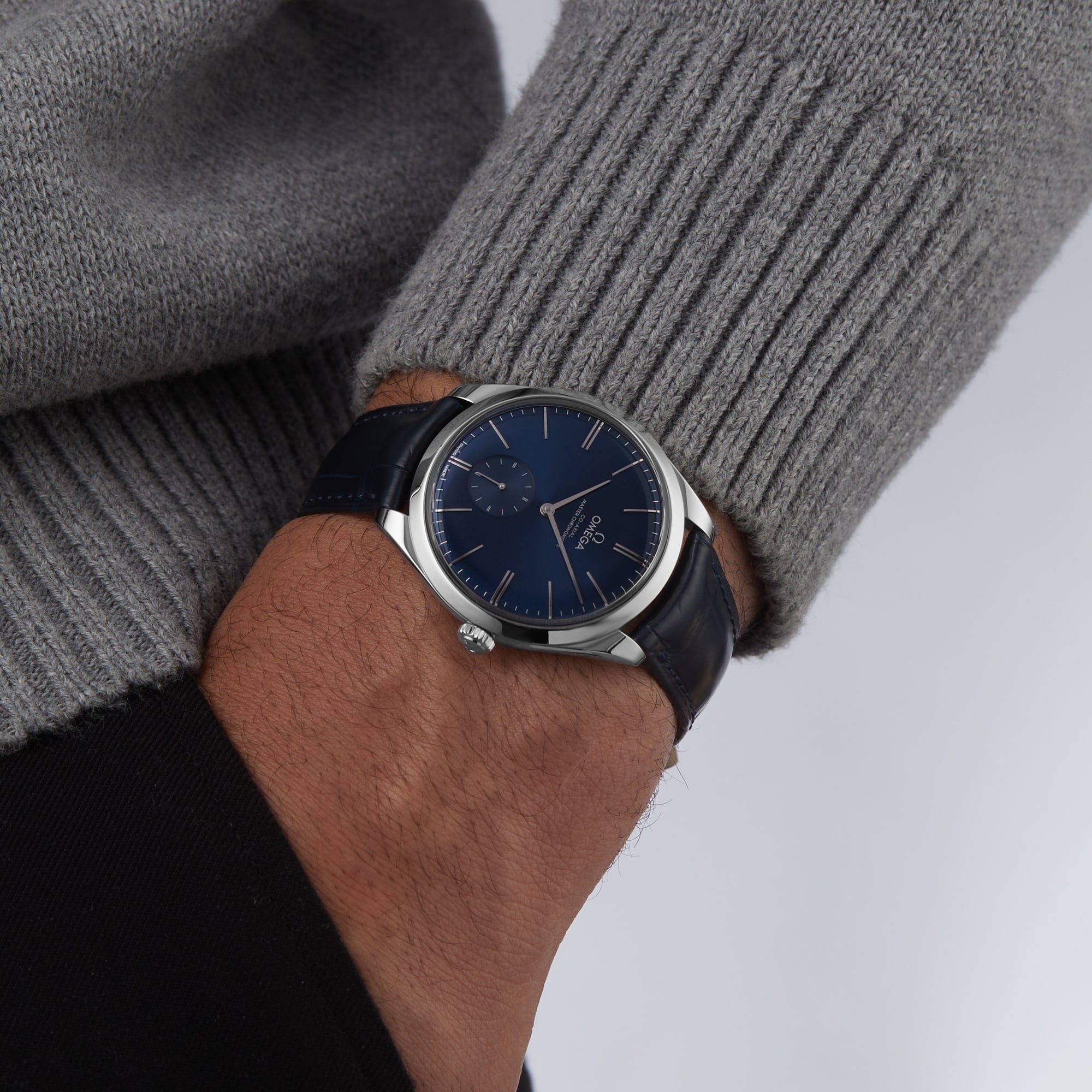 A close-up of a person wearing an OMEGA De Ville Trésor with a blue dial and leather strap, powered by the Master Chronometer-certified Co-Axial Calibre 8926, paired with a gray knitted sweater.