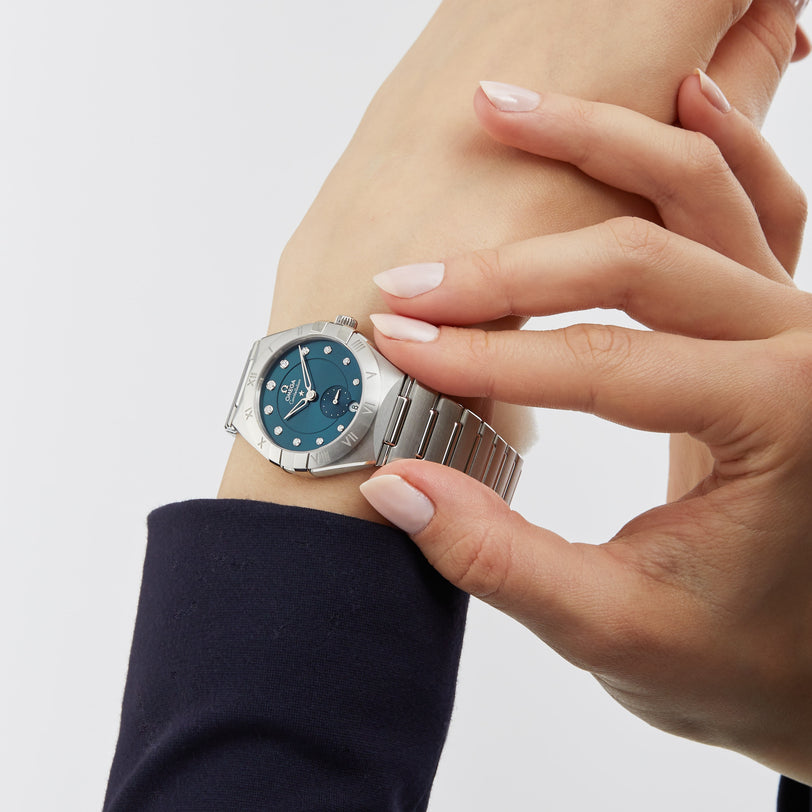 Someone adjusts an OMEGA Constellation 34mm wristwatch, featuring diamond hour markers and a blue dial, on their left wrist against a plain white background.