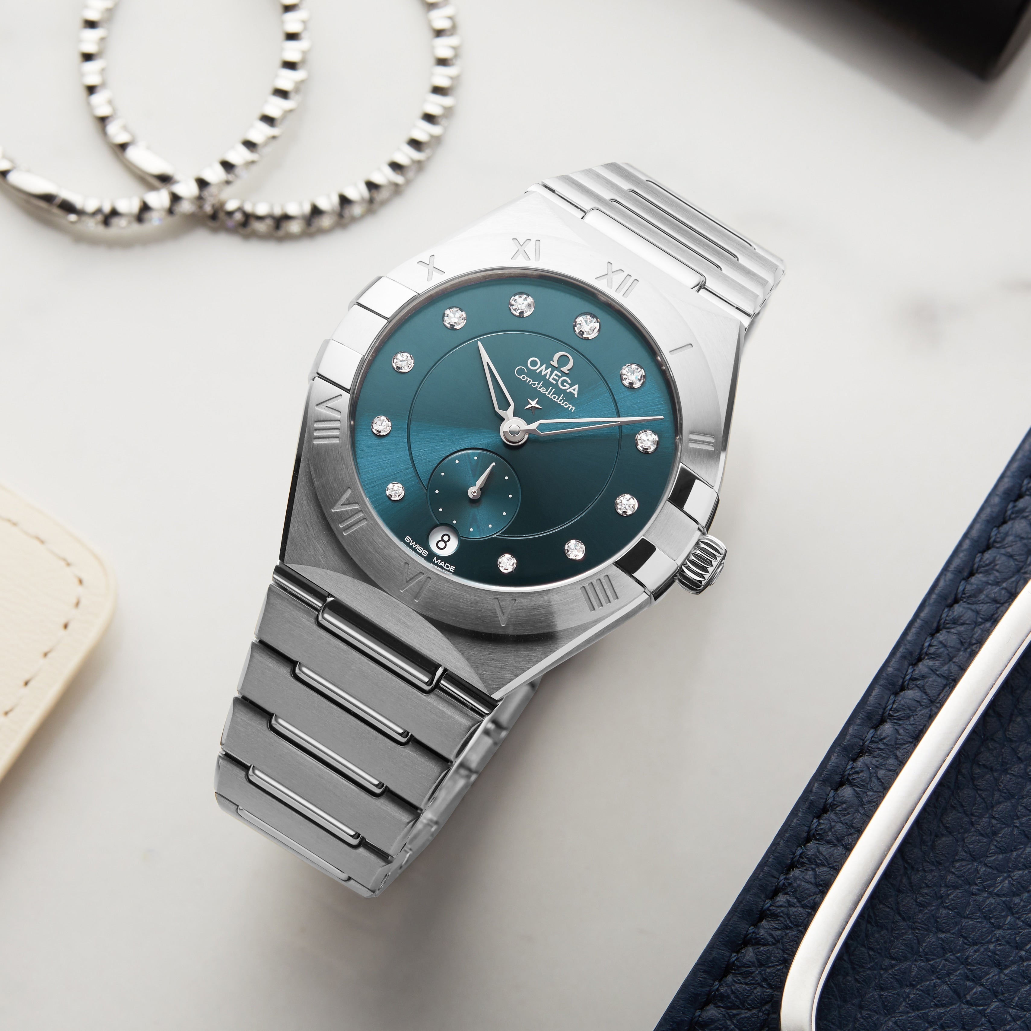 An OMEGA Constellation stainless steel wristwatch with a teal dial, diamond hour markers, and Roman numerals on the bezel is displayed on marble beside jewelry and leather goods.