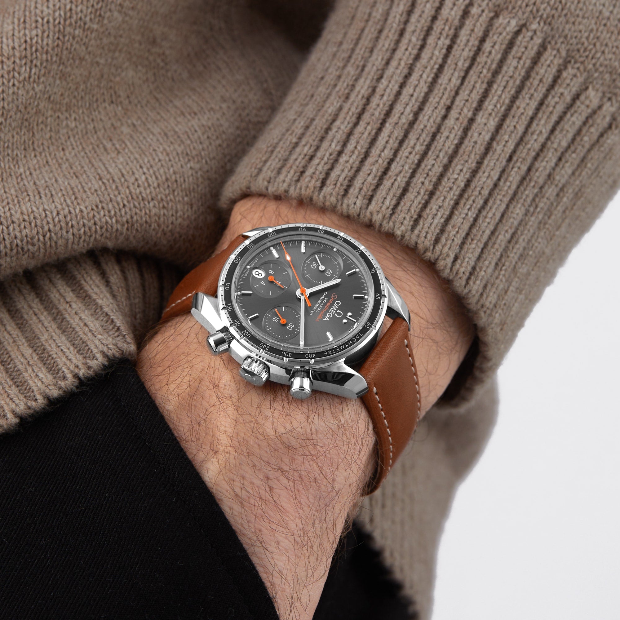 Wearing a beige sweater and black pants, a person sports the OMEGA Speedmaster 38—a sporty, stylish chronograph with a brown leather strap, gray dial, and three sub-dials.