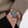 Wearing a beige sweater and black pants, a person sports the OMEGA Speedmaster 38—a sporty, stylish chronograph with a brown leather strap, gray dial, and three sub-dials.