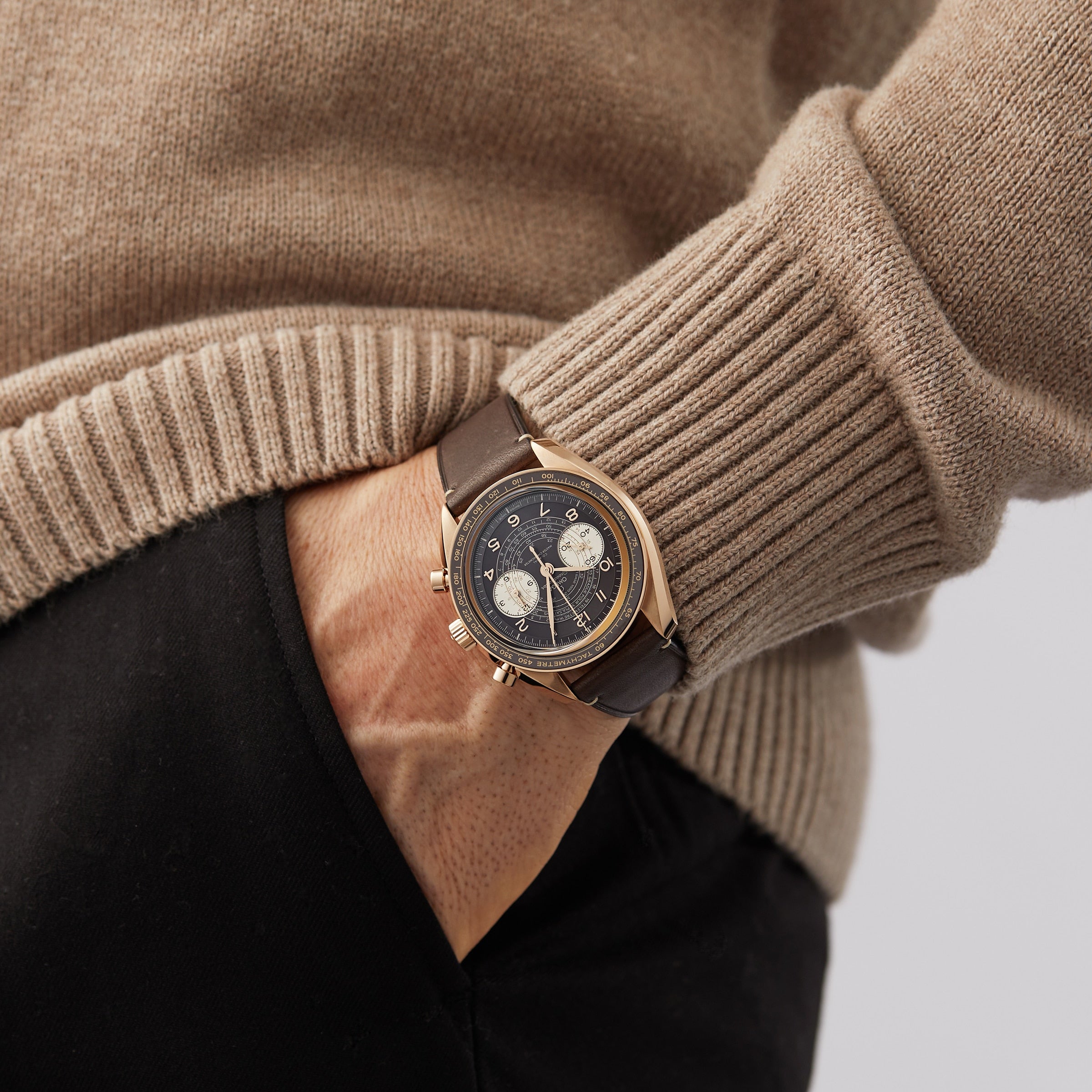 A person in a beige sweater and black pants puts their hand in their pocket, displaying an OMEGA Speedmaster Chronoscope with a gold case, dark dial, and leather strap that highlights its vintage-inspired design.