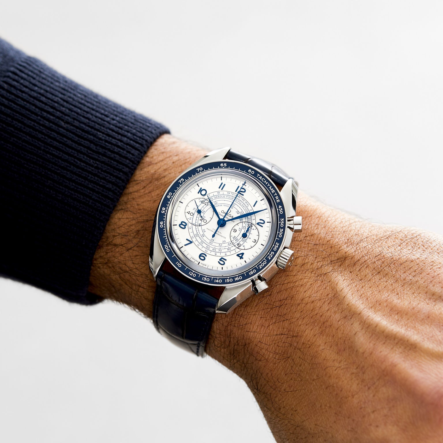 A wrist in a navy long-sleeve shirt wears an OMEGA Speedmaster Chronoscope, a silver watch with a white dial, blue accents, and a blue leather strap—boasting 1940s-inspired design elements.