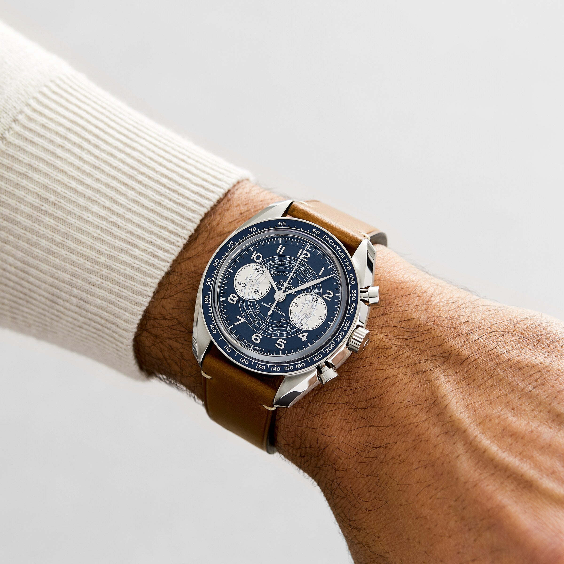 Someone is wearing an OMEGA Speedmaster Chronoscope with a blue dial, 9908 calibre movement, brown leather strap, and a beige long sleeve sweater.