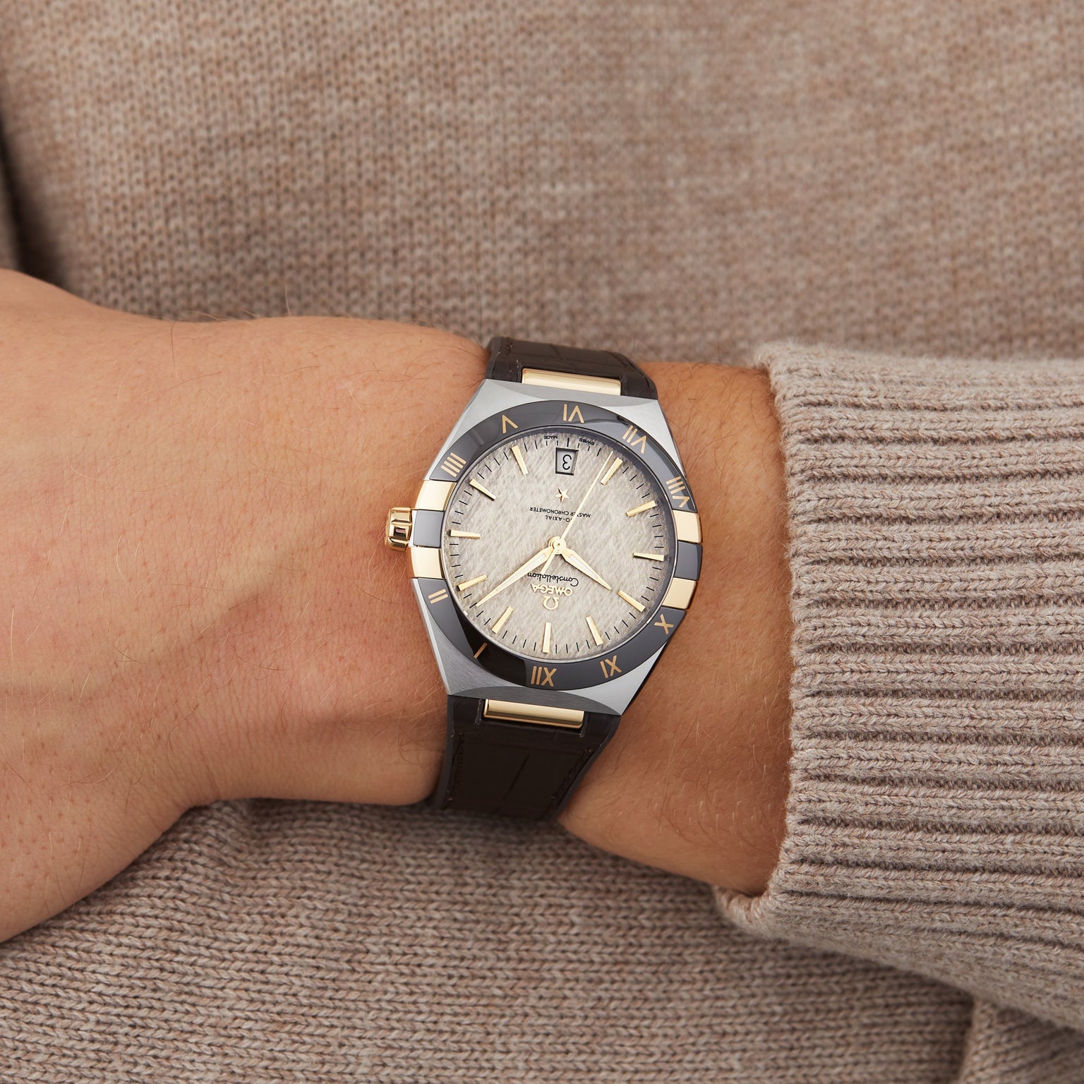 A person wears the OMEGA Constellation, a gold and silver wristwatch with a leather strap, Roman numerals, and master chronometer certified 8900 calibre movement on their left wrist over a beige knit sweater.