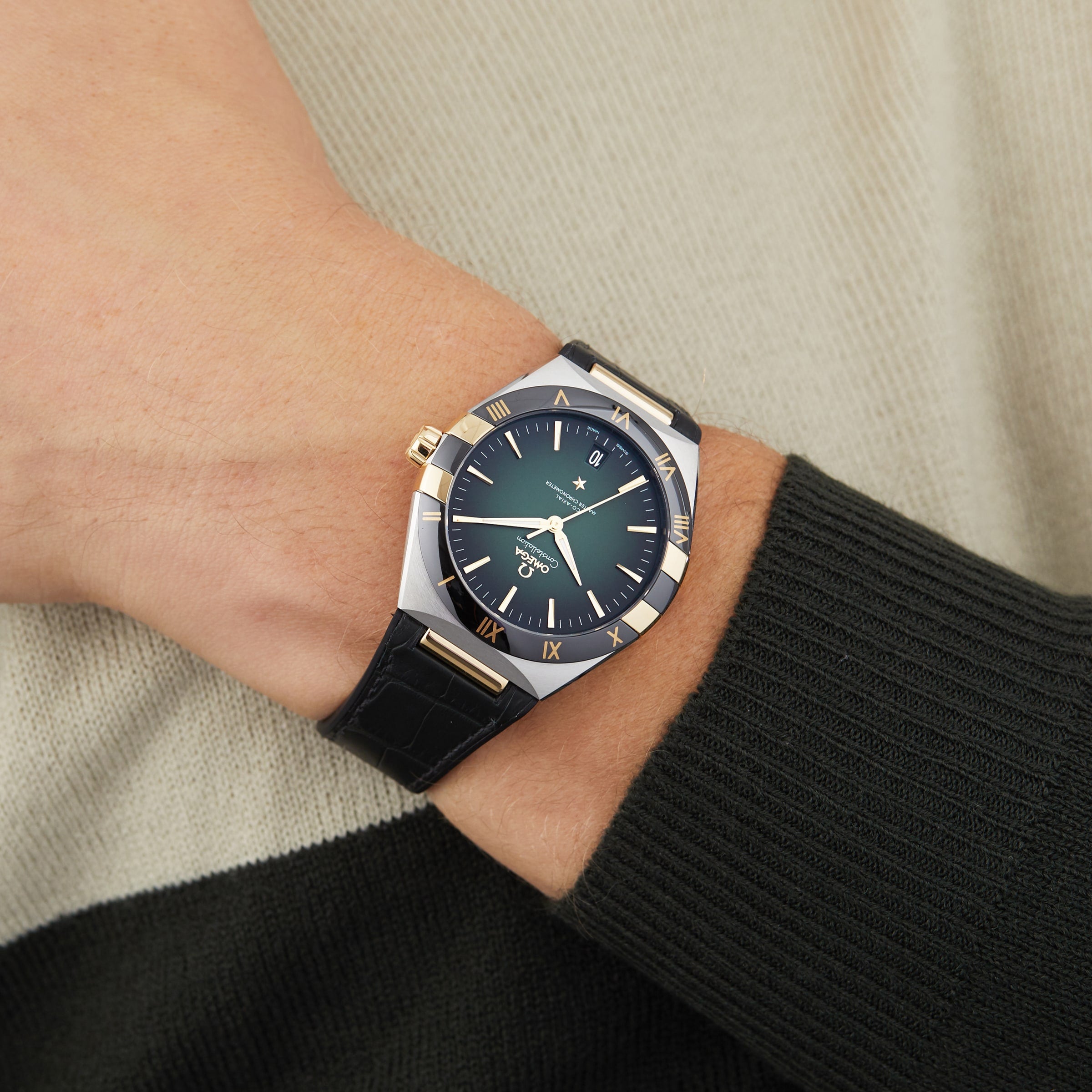 An OMEGA Constellation with a green dial, gold Roman numerals, and black leather strap is worn over a green and white sweater. This elegant watch is master chronometer certified and powered by the 8900 calibre movement.