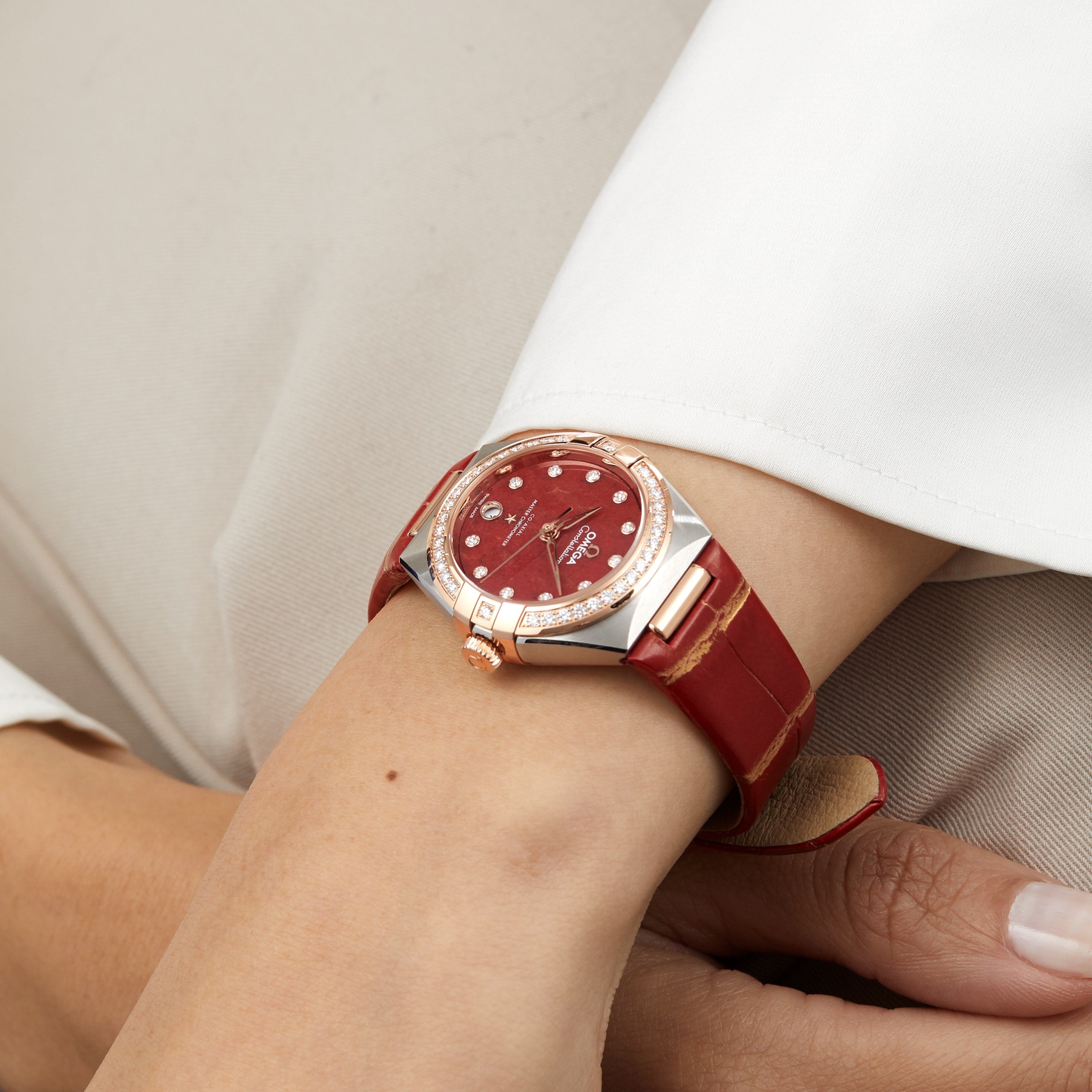 A person wears an OMEGA Constellation wristwatch with a red leather strap, red face, and gold accents—an eye-catching timepiece master chronometer certified for precision.