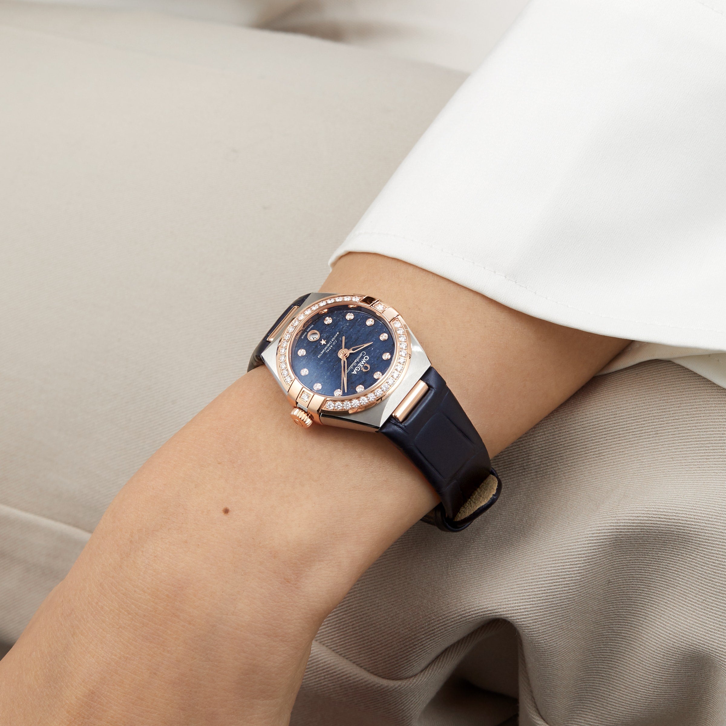 A close-up of a person wearing an OMEGA Constellation watch with an aventurine dial, 18K Sedna™ gold accents, and a black band, resting their arm on their lap.