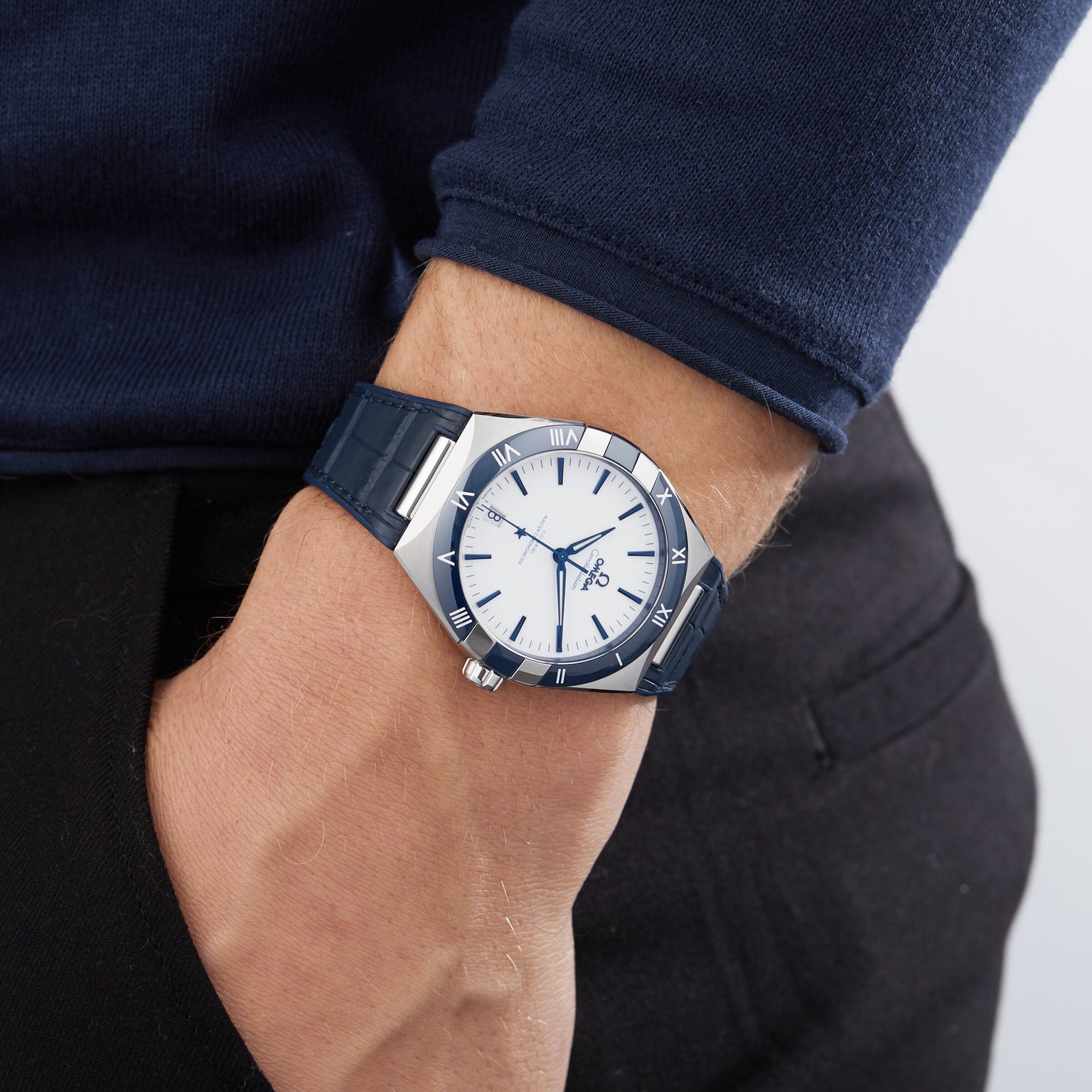 A person in a dark blue long-sleeve shirt and black pants wears an OMEGA Constellation on their left wrist, their hand casually tucked in their pocket.