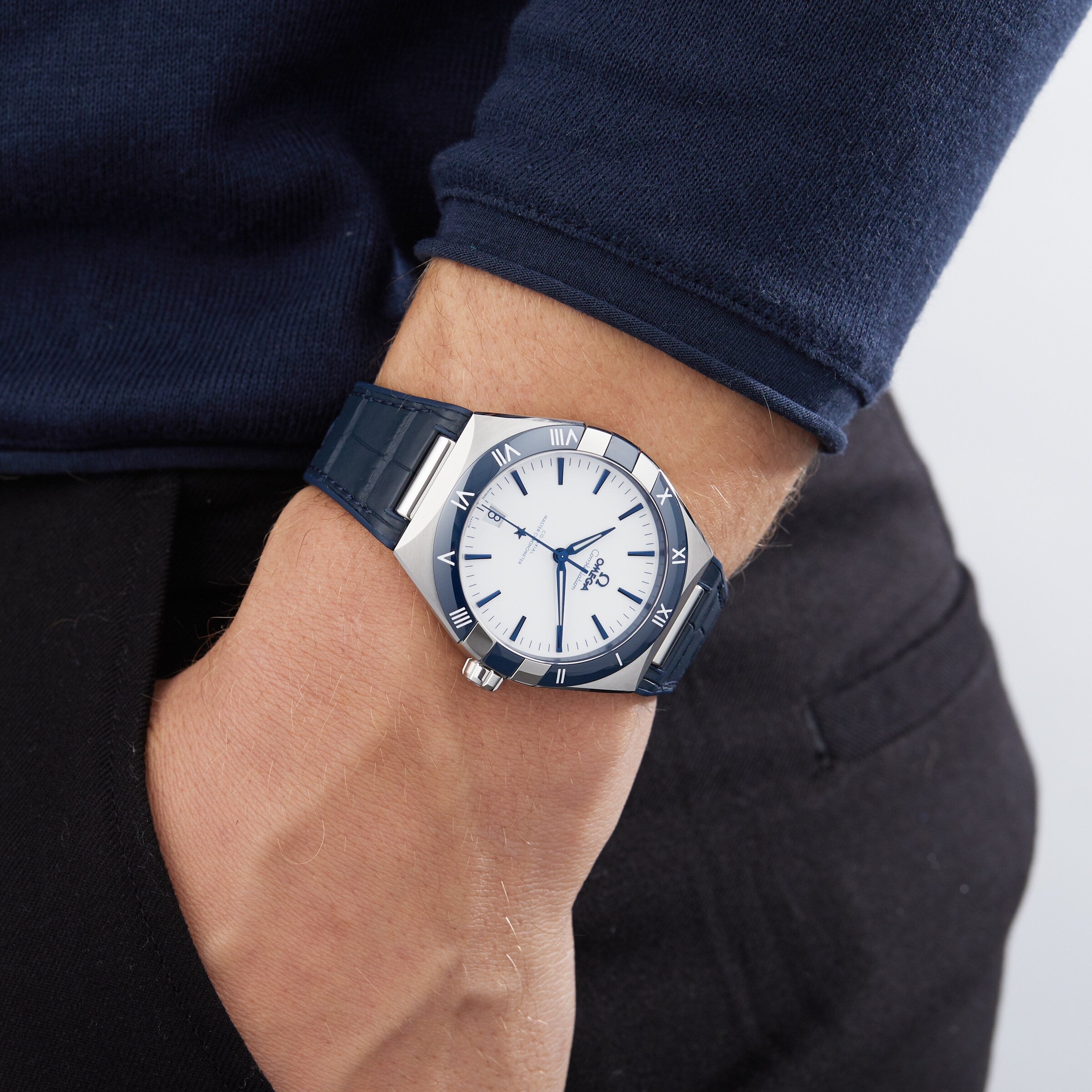 A person in a dark blue long-sleeve shirt and black pants wears an OMEGA Constellation on their left wrist, their hand casually tucked in their pocket.