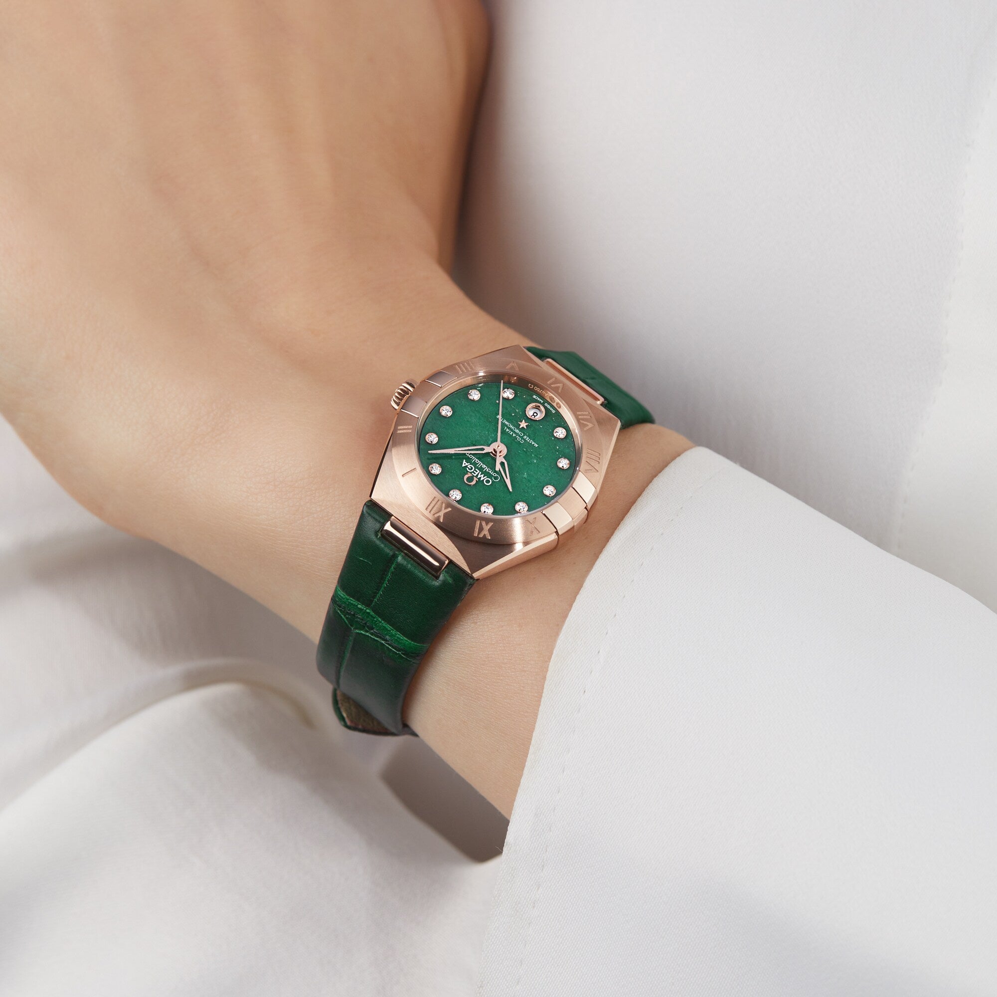 A person wears the OMEGA Constellation, featuring a green face, green leather strap, and 18K Sedna™ gold case, shown against a white sleeve.