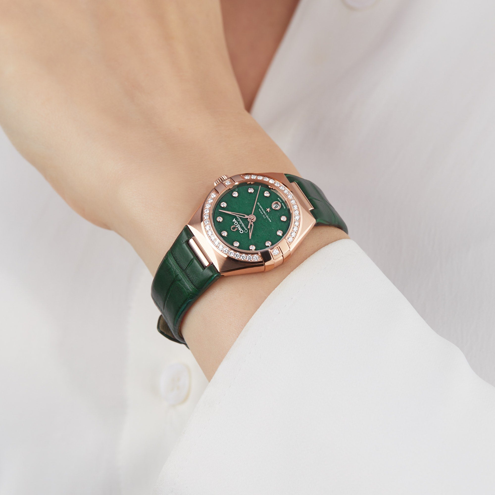 A person wearing the OMEGA Constellation, featuring an aventurine green face, diamond accents, Sedna™ gold case, and green leather strap, styled with a white long-sleeve shirt—showcasing elegant sophistication.