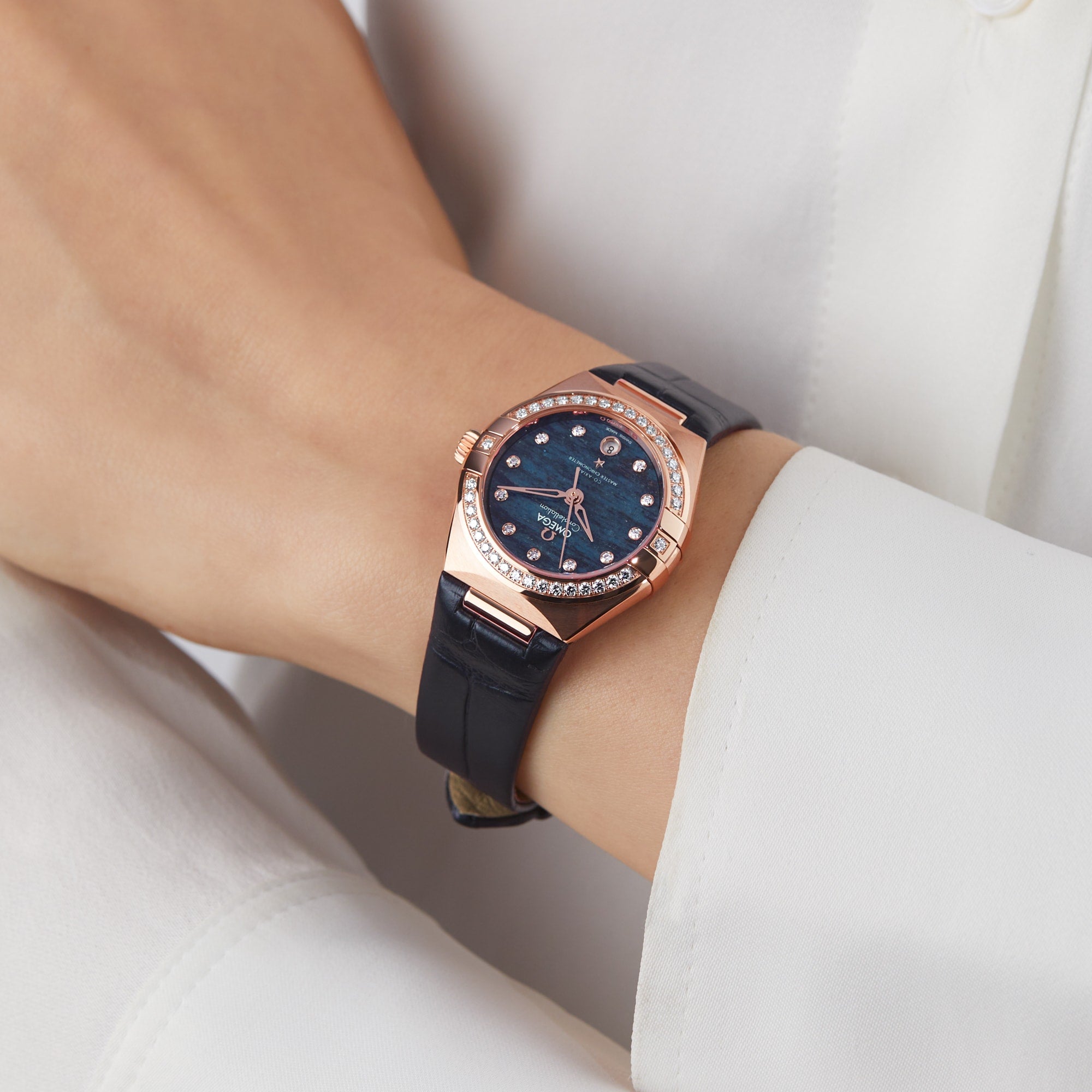 A person wears an OMEGA Constellation with an 18K Sedna™ gold case, a deep blue Aventurine dial set with diamond markers, and a black leather strap, displayed on their left wrist.