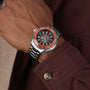 A person in a maroon shirt showcases the OMEGA Seamaster Planet Ocean 6000M—an Ultra Deep stainless steel wristwatch with a black dial, orange bezel, and O-MEGASTEEL construction designed for saturation divers.