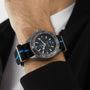 A close-up of a wrist wearing an OMEGA Seamaster Planet Ocean 6000M dive watch with a black and blue striped nylon strap, partially covered by a black jacket sleeve.