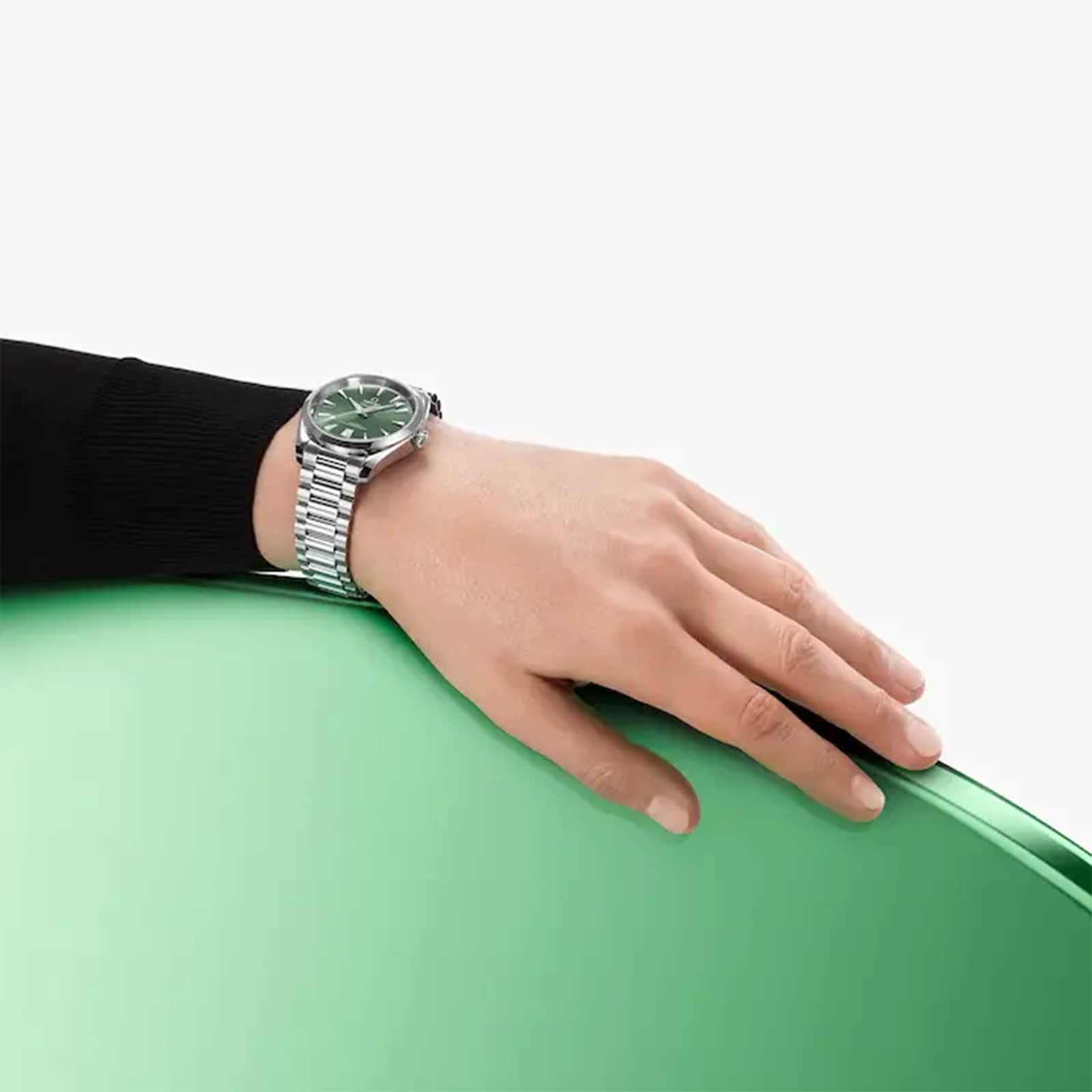 A hand wearing an OMEGA Speedmaster 57 with a green dial and Master Chronometer certification rests on a smooth, rounded green surface.