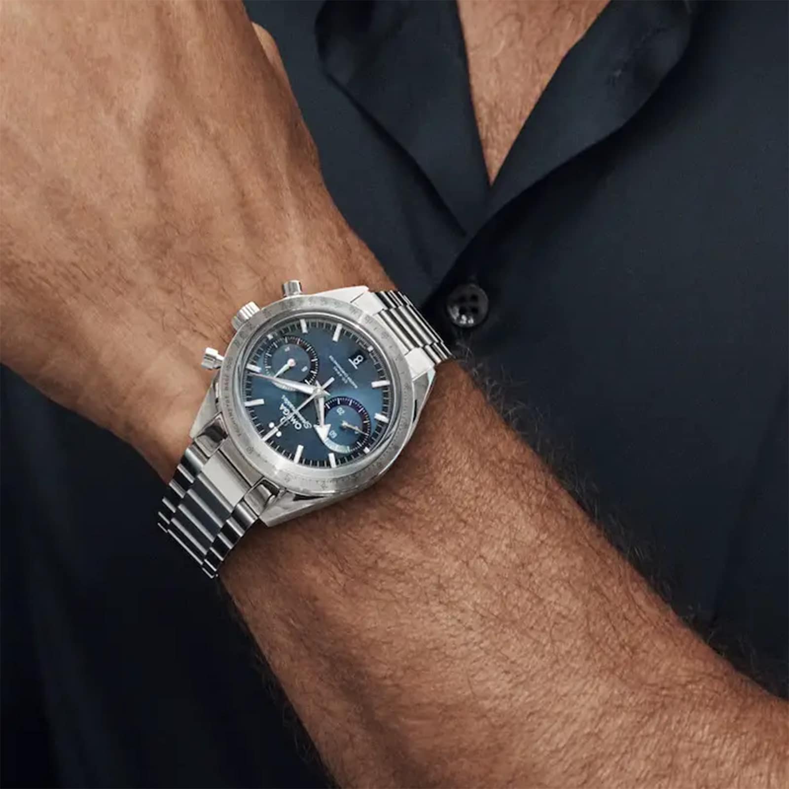 A person in a short-sleeve black shirt wears the OMEGA Speedmaster 57 chronograph, featuring calibre 9906, a blue dial, and a metal strap.