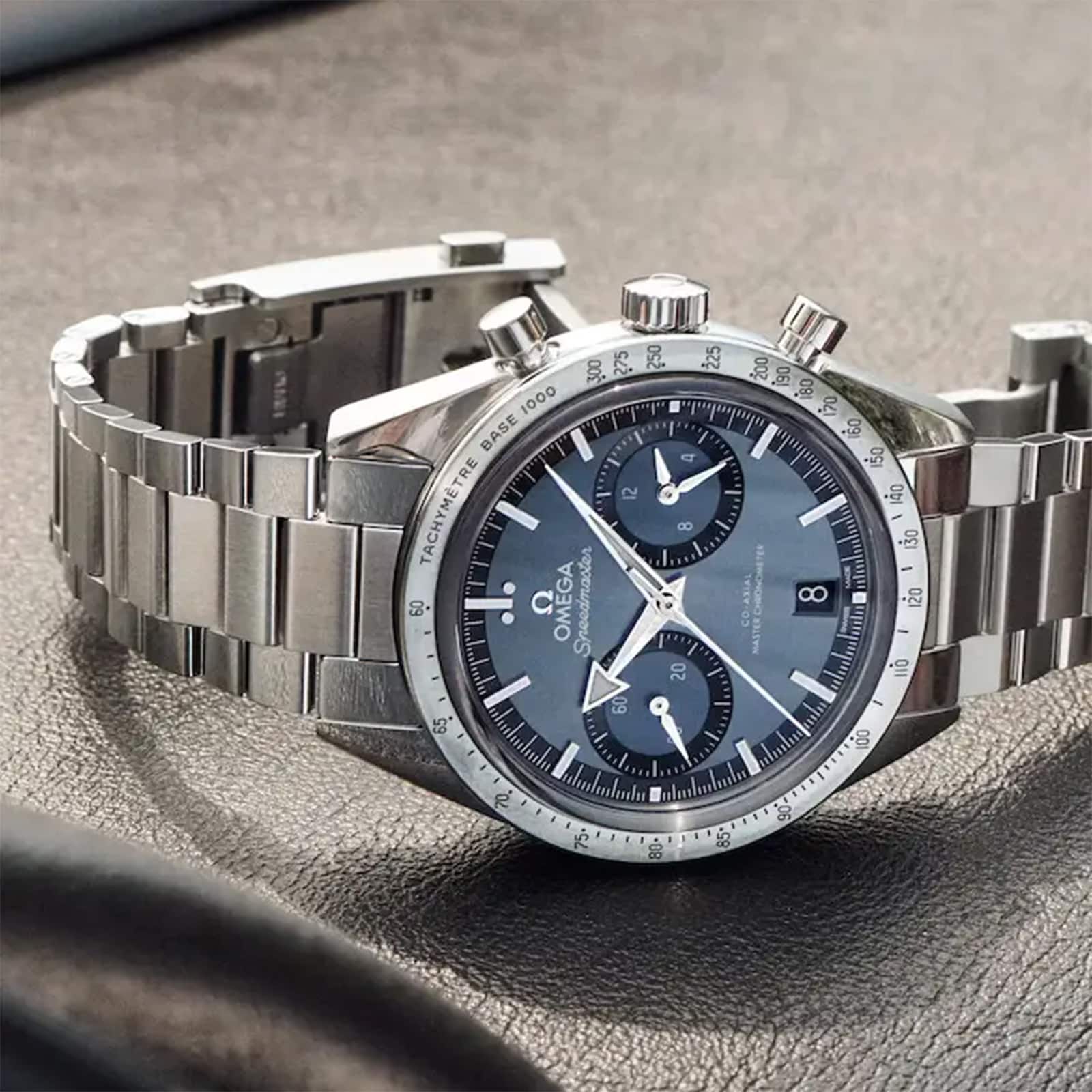 An OMEGA Speedmaster 57 chronograph wristwatch with a blue dial and two subdials, featuring calibre 9906 movement, is displayed on a textured brown surface.