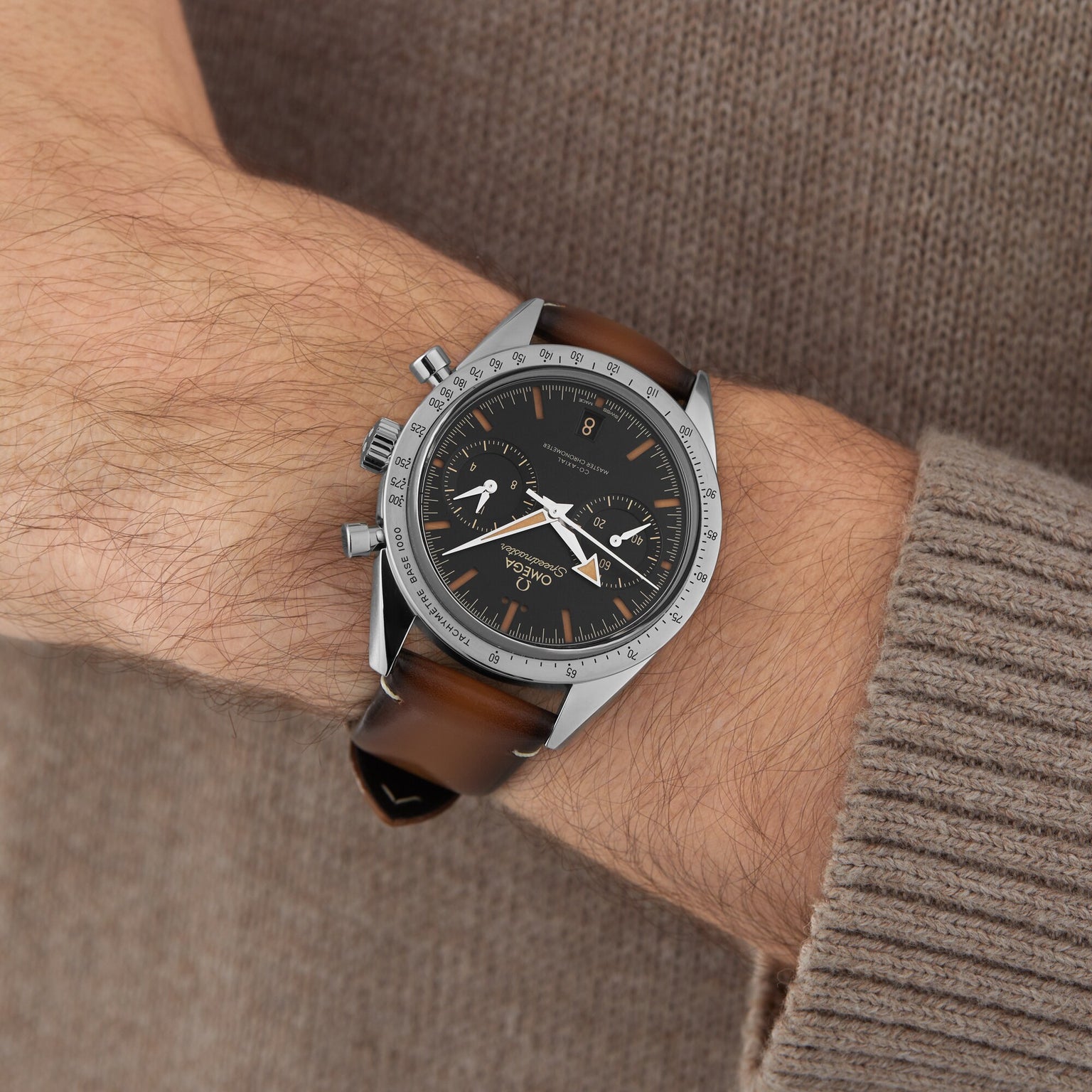A close-up of a wrist wearing an OMEGA Speedmaster 57 with a brown leather strap, black dial, silver case, and Calibre 9906 movement over the sleeve of a brown knit sweater.
